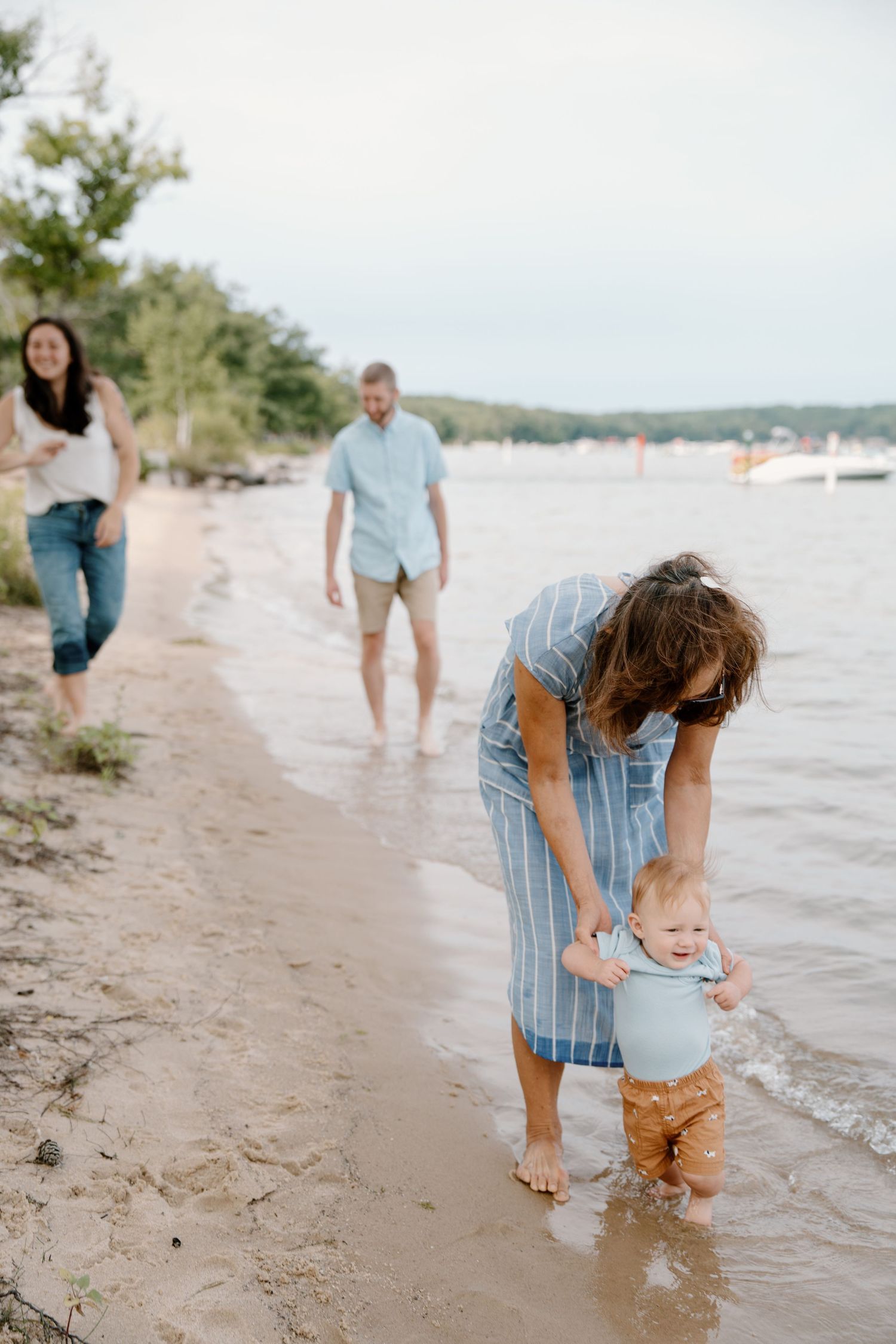 Family enjoying a beach day, mother helping toddler walk in shallow water while father and woman walk along sandy shore.