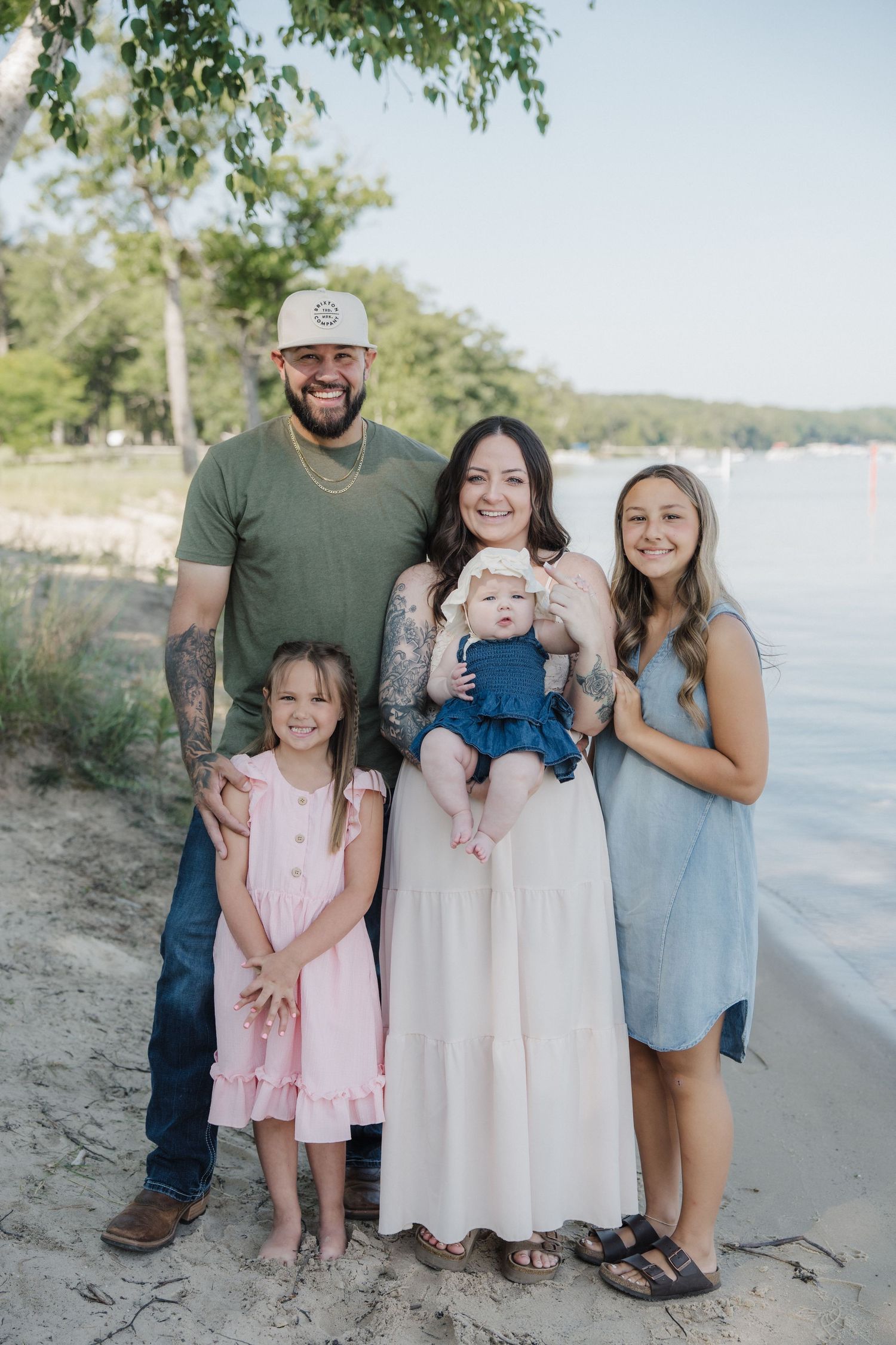 Family of five posing on a sandy beach shore, parents with three daughters, smiling in casual summer outfits near a lake.