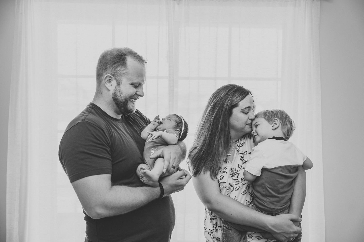 Black and white family portrait of parents holding newborn and toddler near bright window.