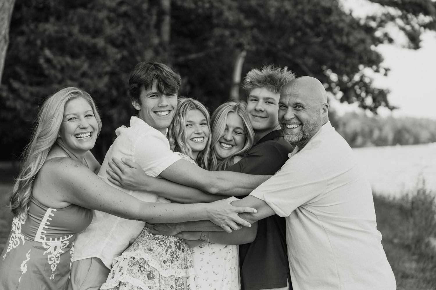Black and white family portrait of six smiling people hugging outdoors near trees and a lake.