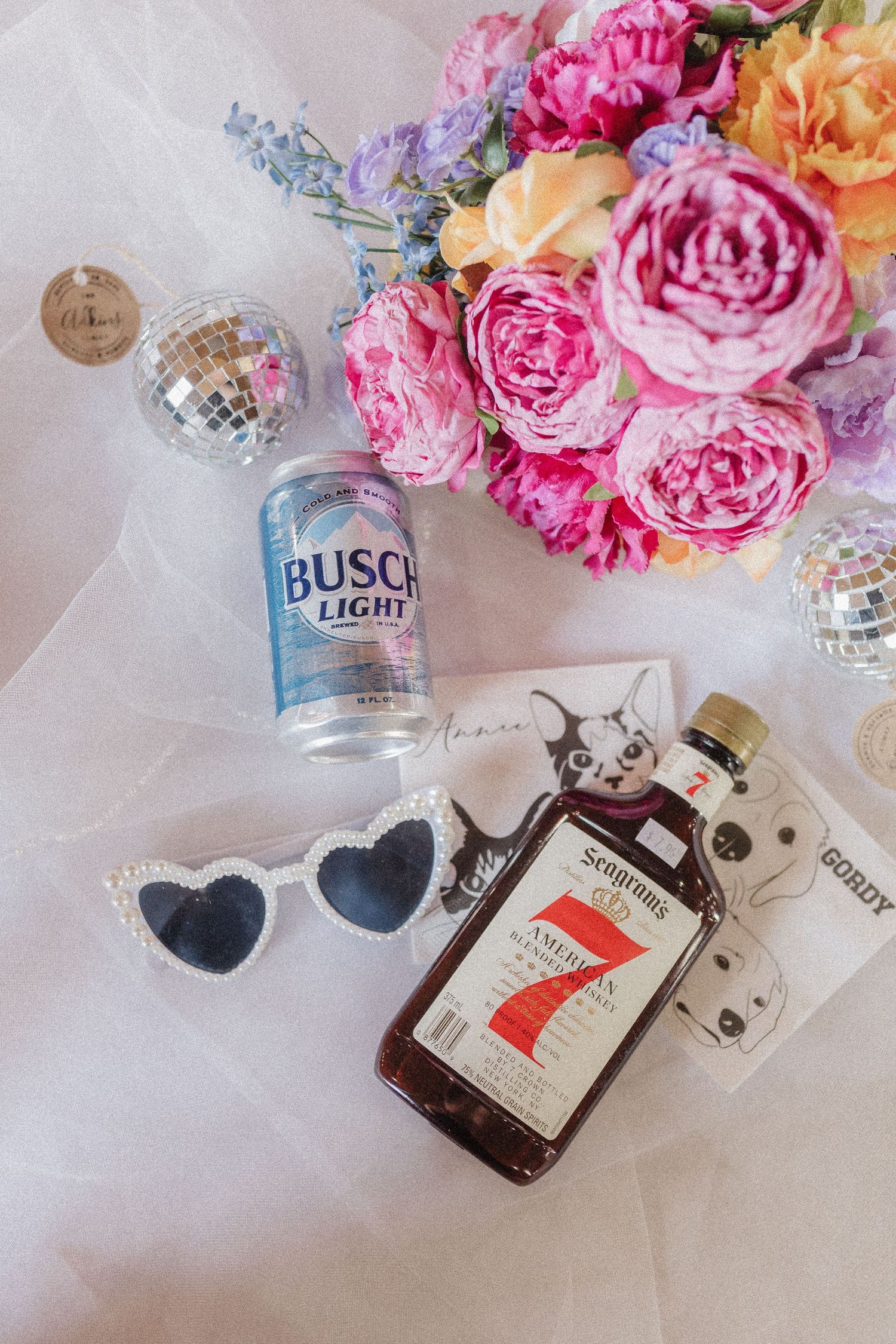 Wedding detail flatlay with pink roses, heart sunglasses, and miniature alcohol bottles on white background.