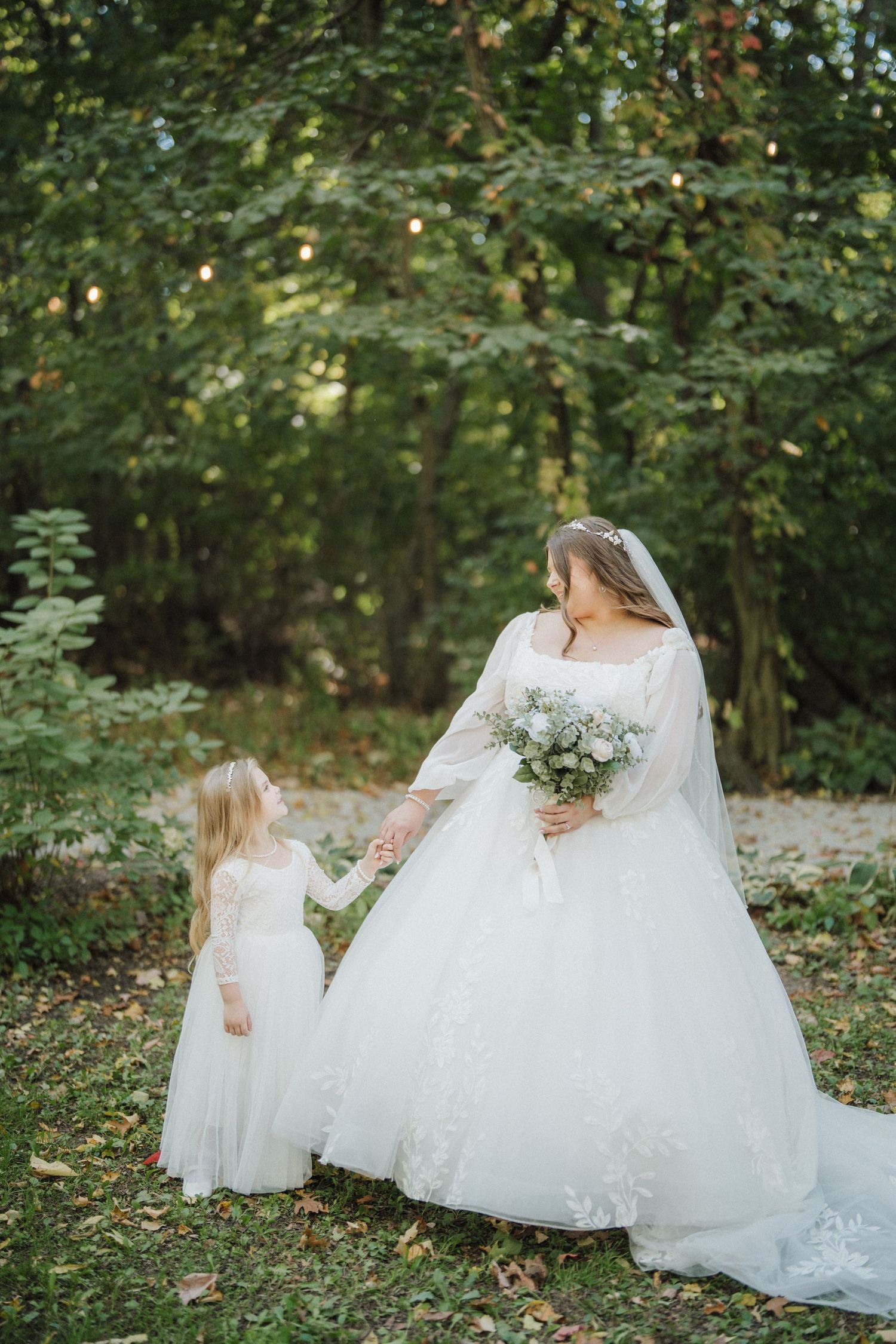 A bride in a flowing white gown holds hands with a small flower girl in matching white dress in a wooded outdoor setting.