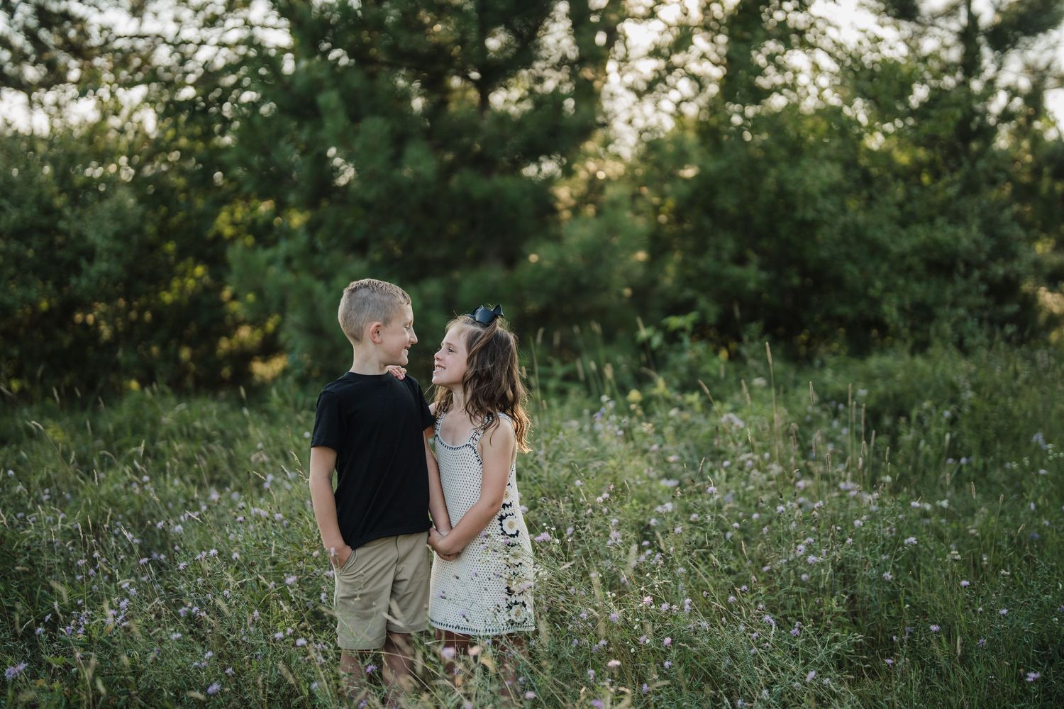 Two people embrace in a romantic pose amidst a field of white wildflowers during a summer evening.