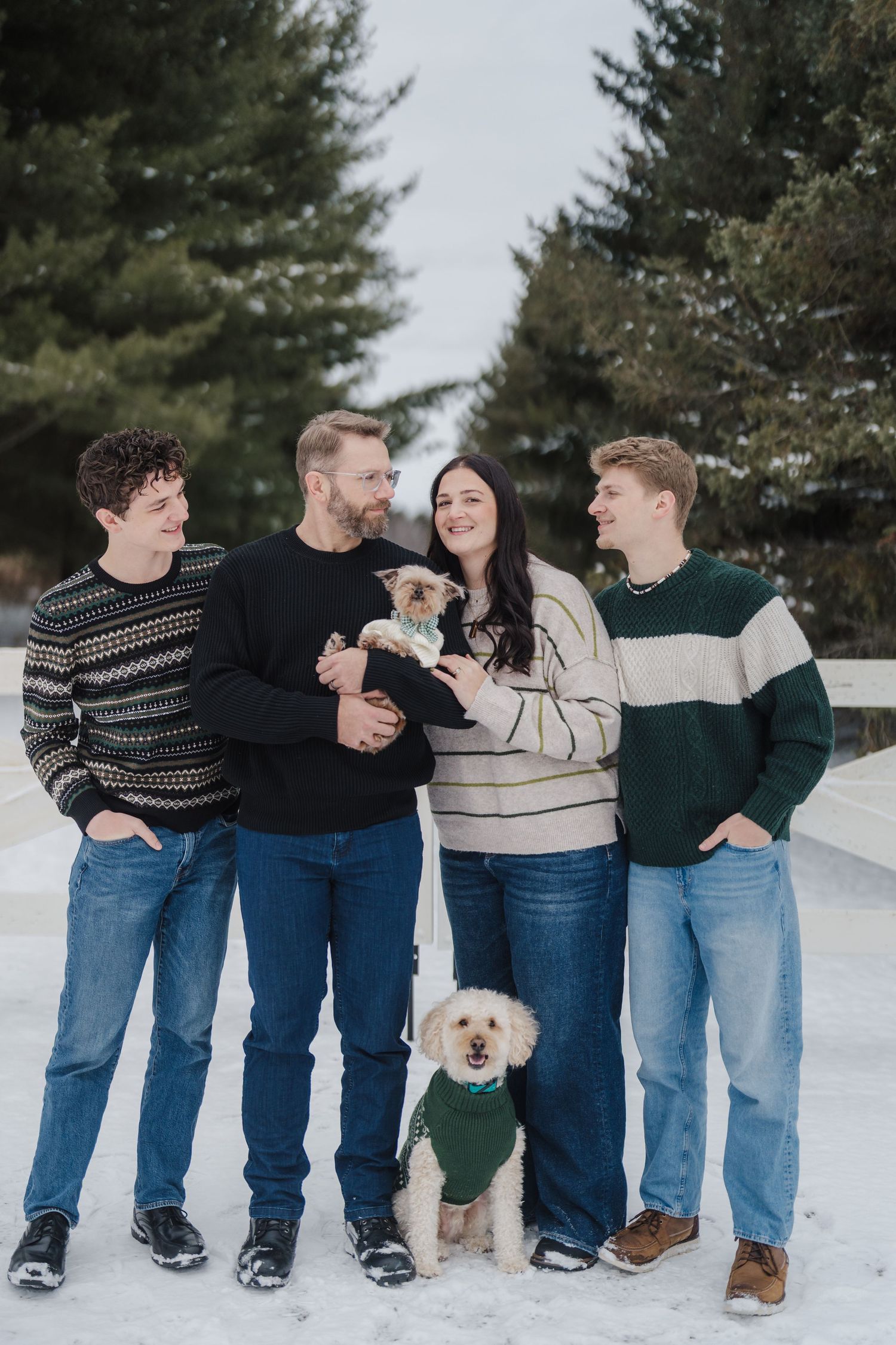 Winter family portrait outdoors in snow with two small dogs wearing matching green sweaters beside evergreen trees.