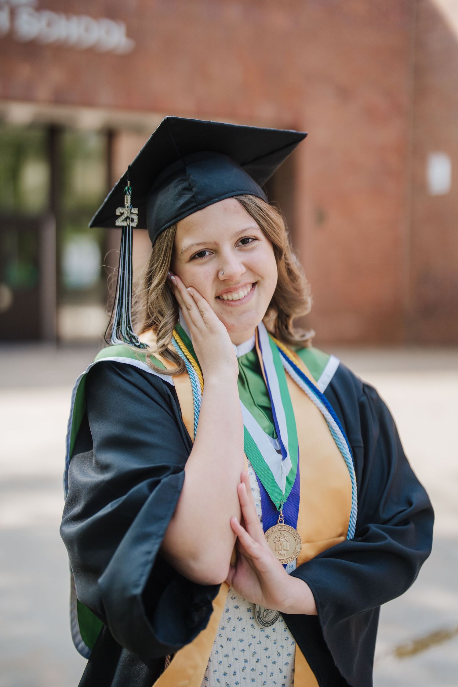 Graduate in cap and gown wearing honor cords and medallion poses outdoors against brick building.