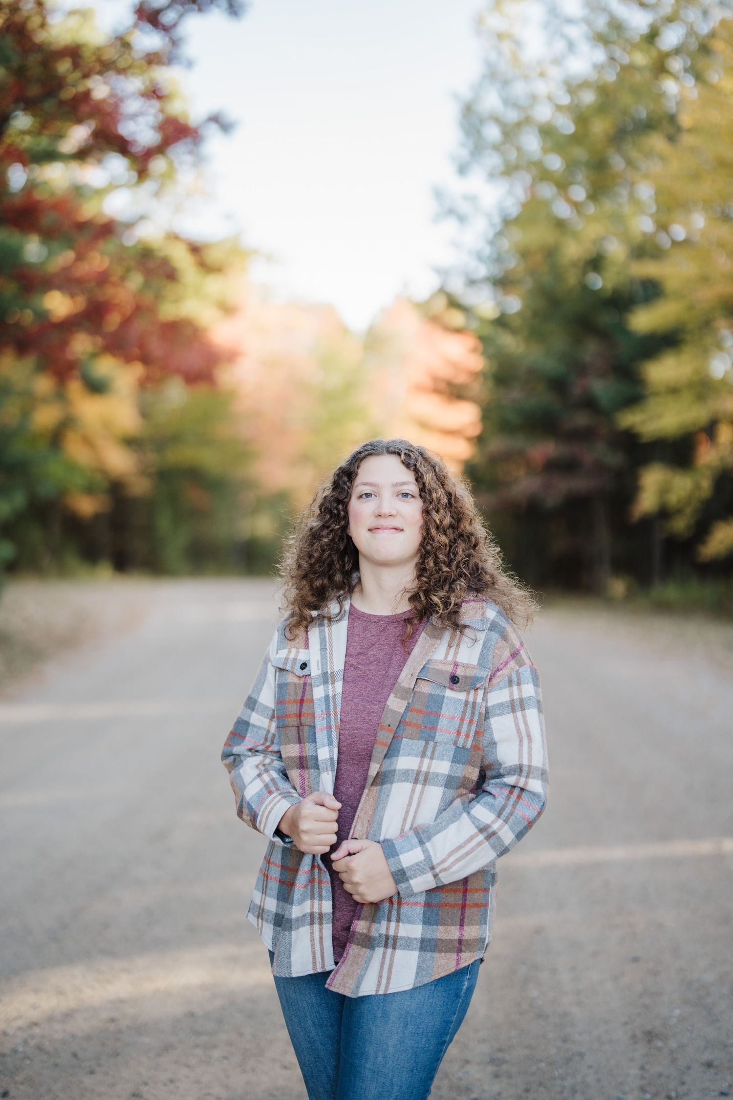 A person wearing a plaid jacket and burgundy top poses outdoors against a backdrop of autumn trees.