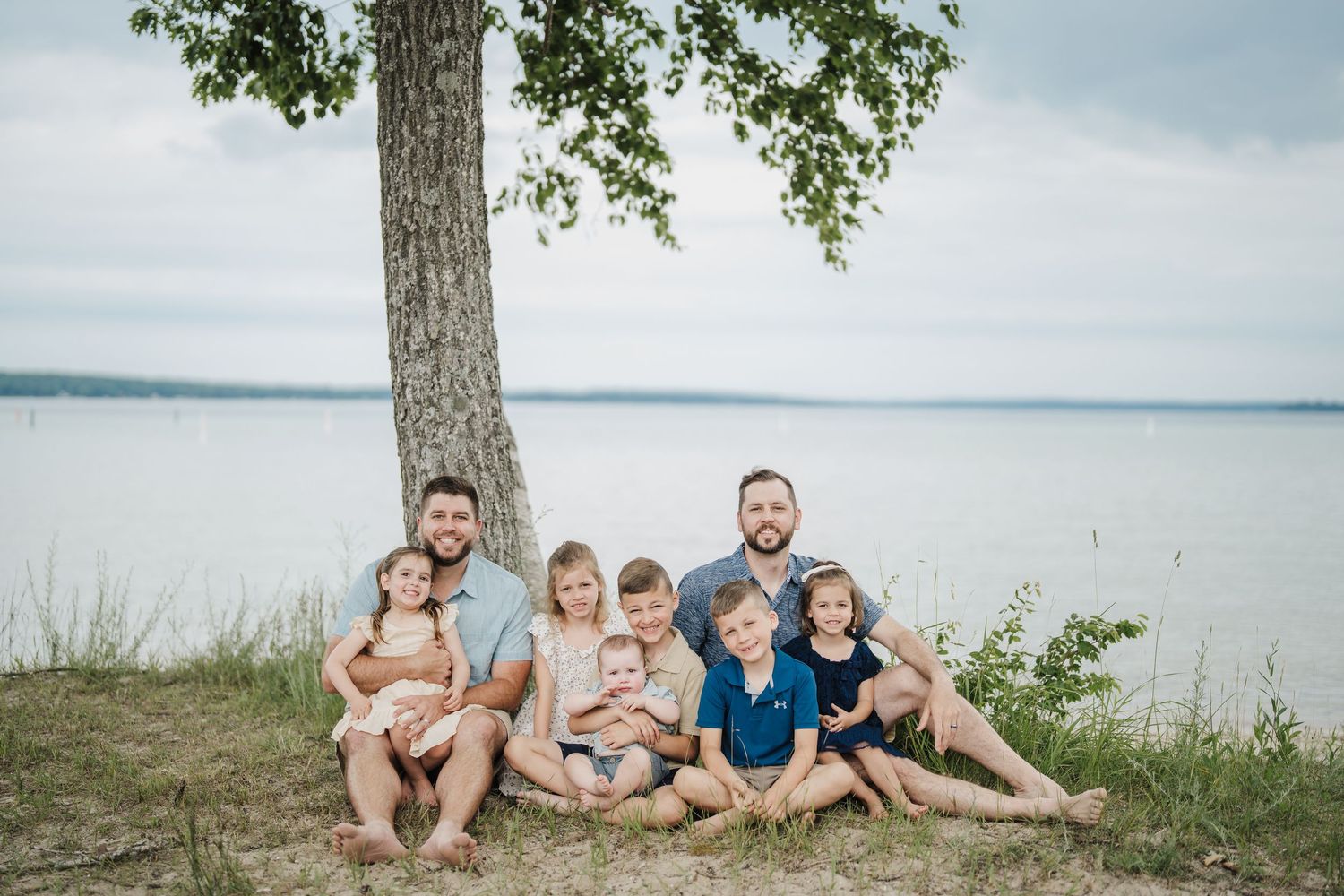 Family group sits together under large tree by peaceful lake shore on cloudy summer day.