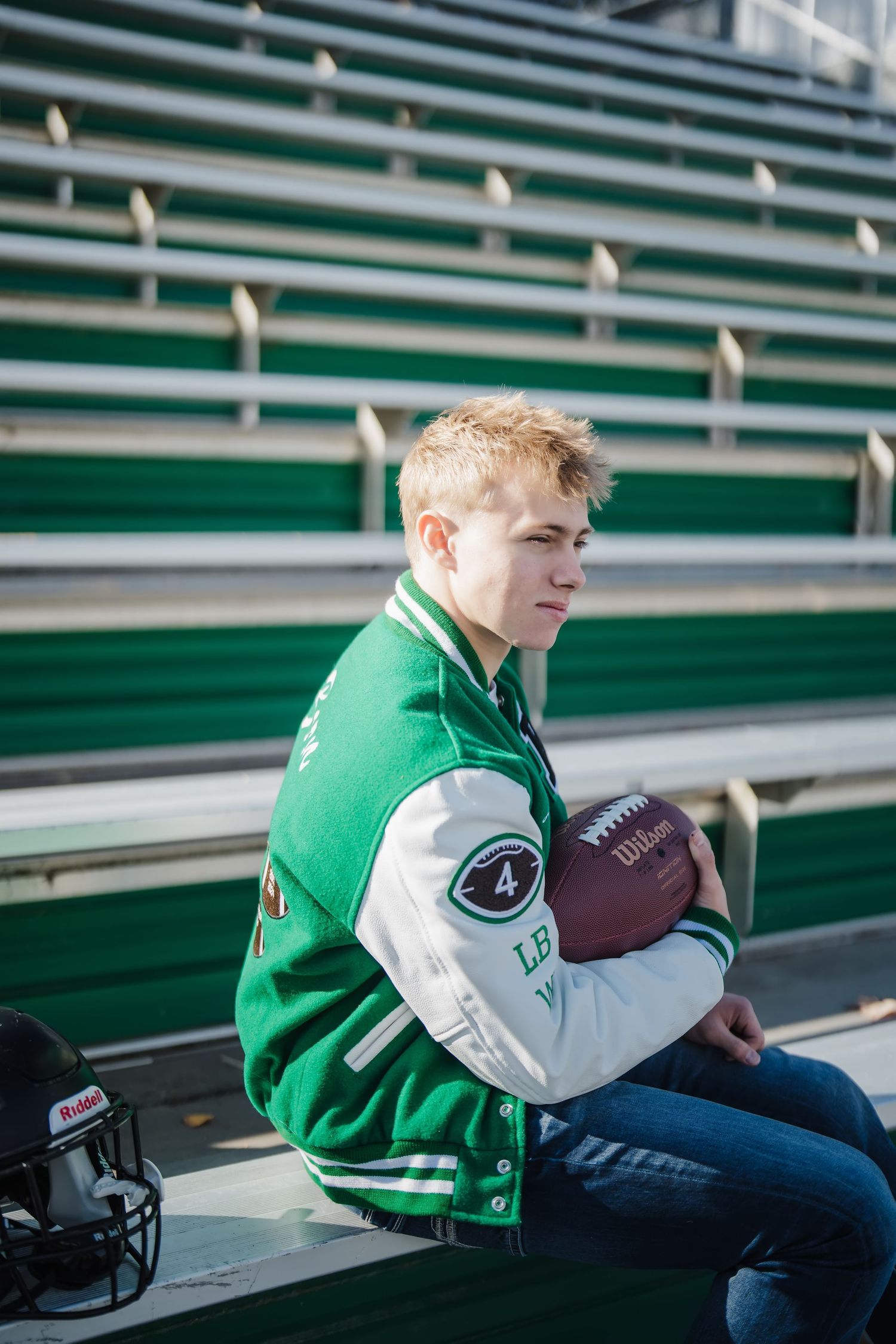 Football player in green and white letterman jacket sits on stadium bleachers with football.