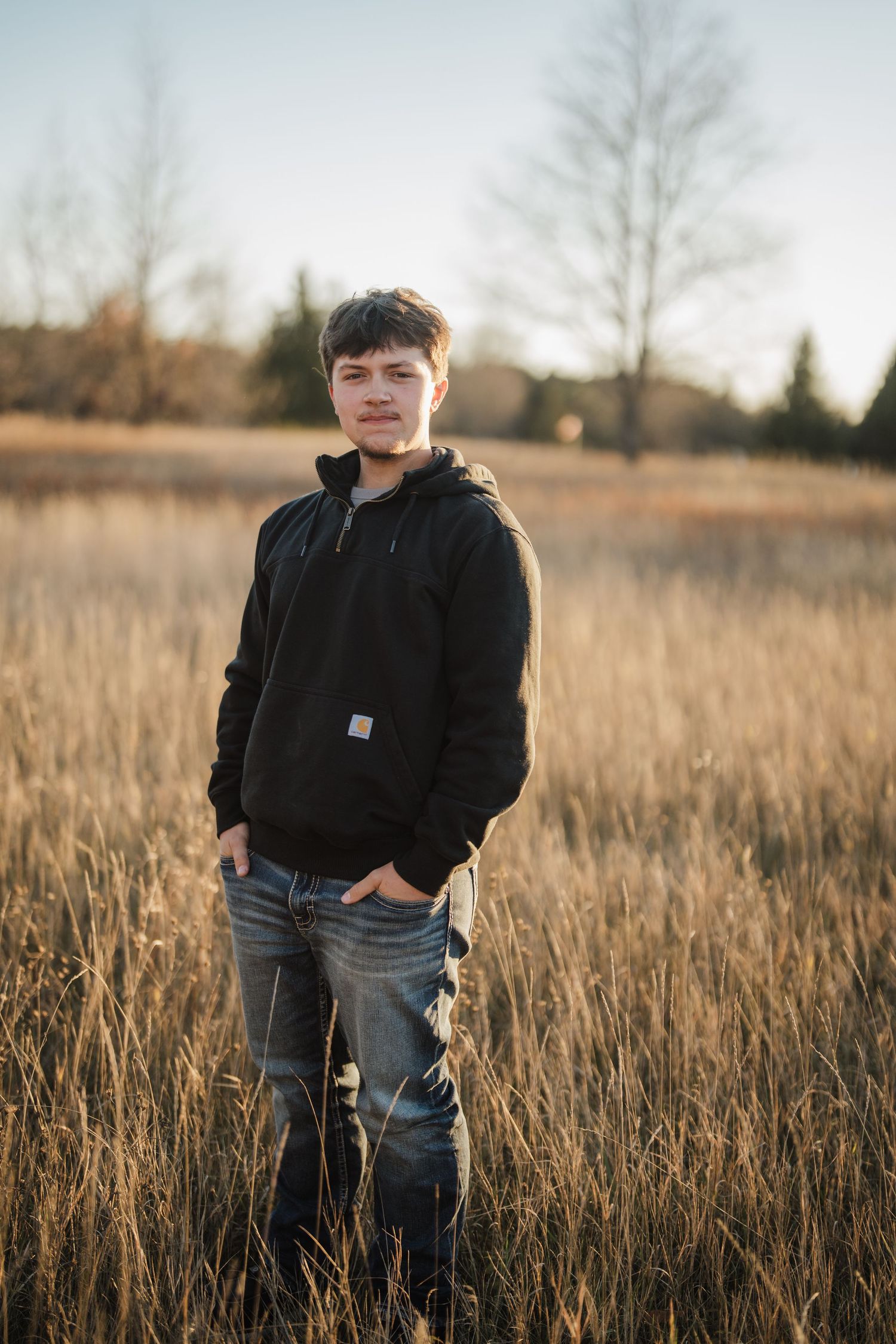 A figure stands in a golden field of tall grass at sunset wearing a black jacket and jeans.