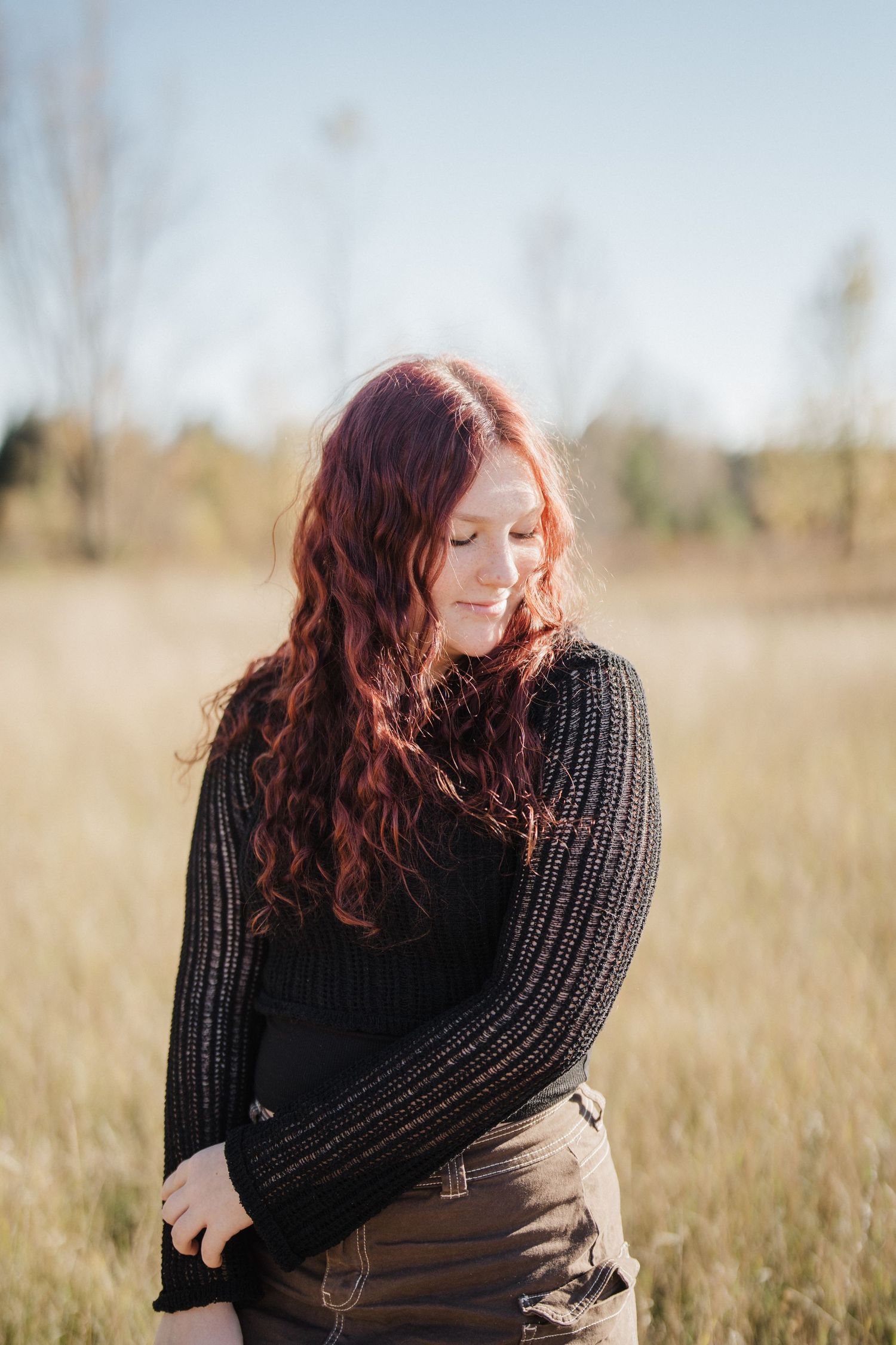 A person with flowing red hair wearing a black sweater poses thoughtfully in a sunlit autumn field.