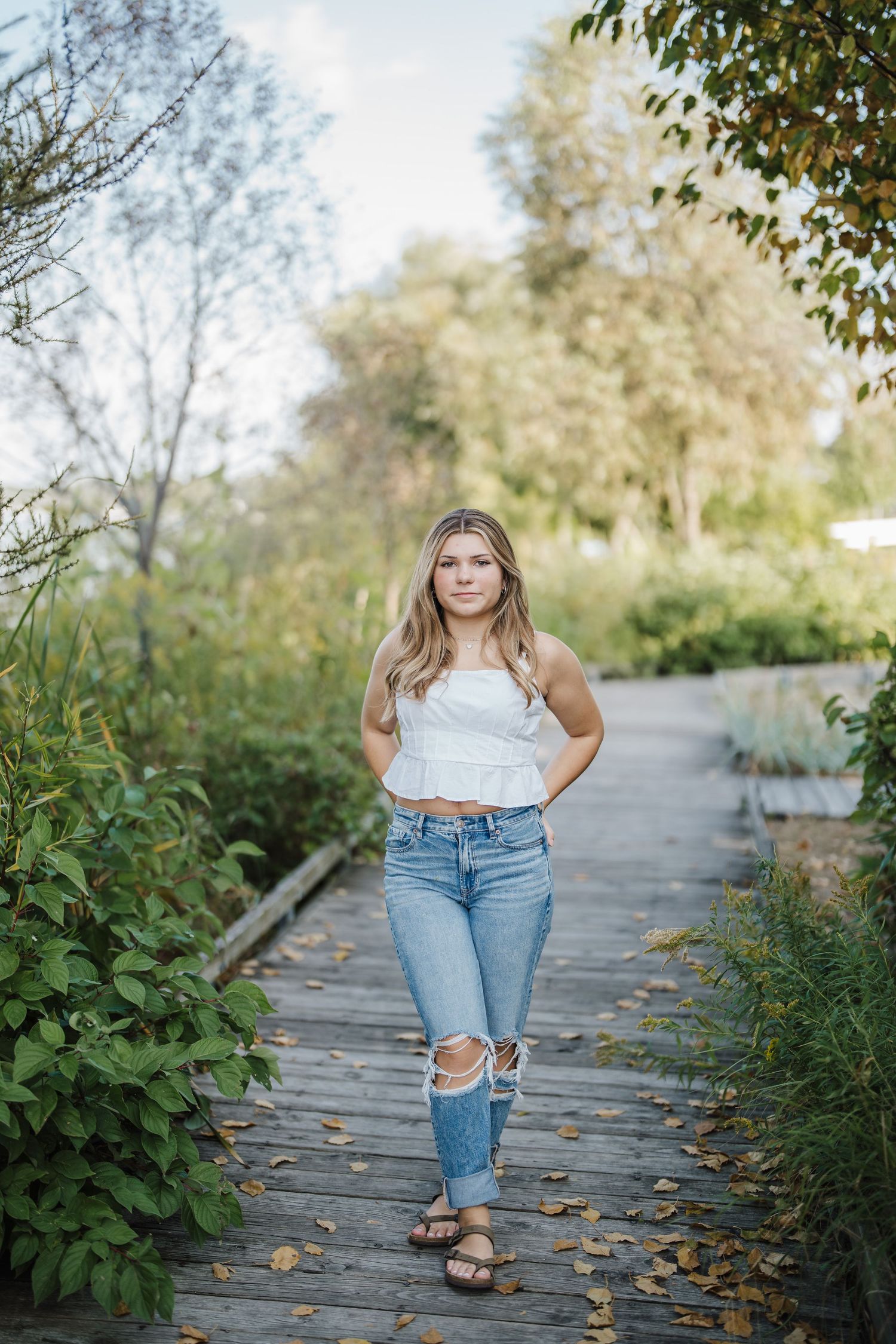 A person in ripped jeans and a white top poses casually on a wooden boardwalk surrounded by autumn foliage.