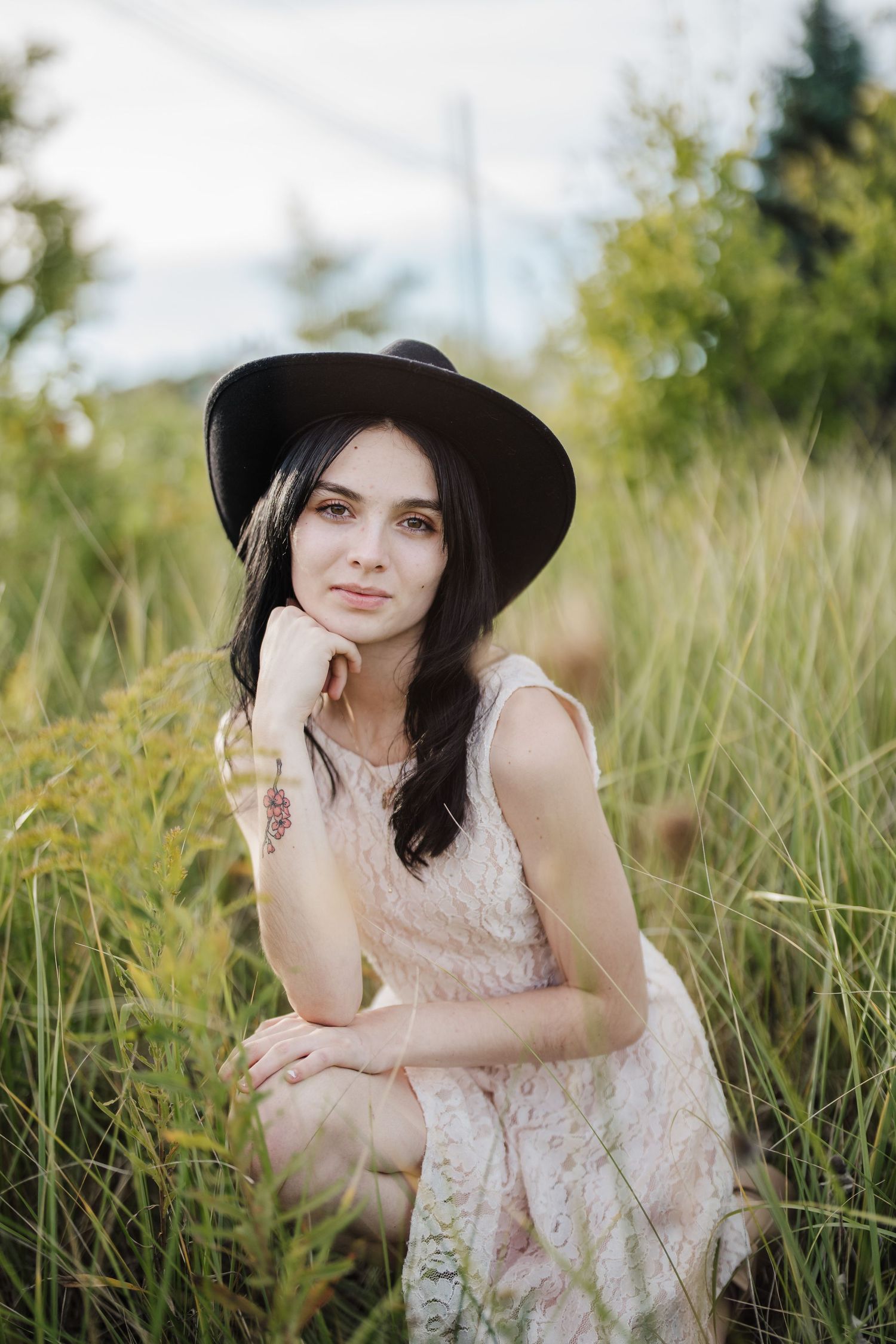 A woman in a floral dress and wide-brimmed hat poses thoughtfully in a sunlit meadow of tall grass.