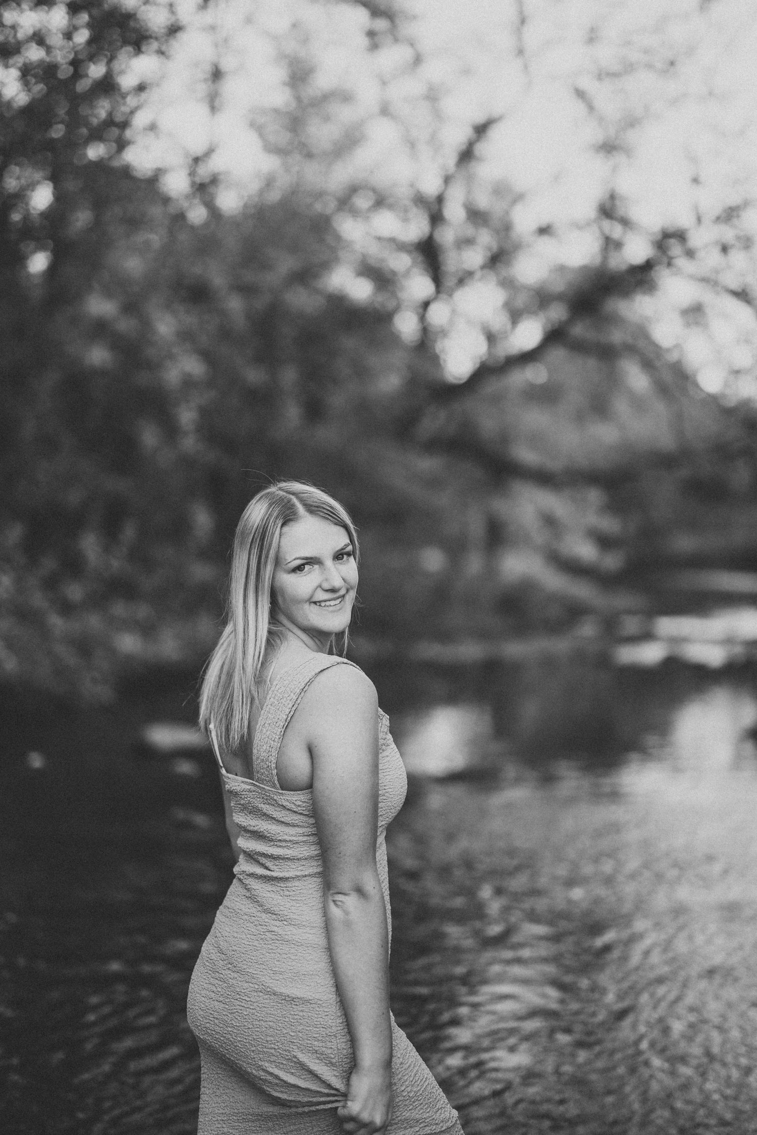 Black and white maternity photo session showing a woman in a sleeveless dress standing by a serene lake.