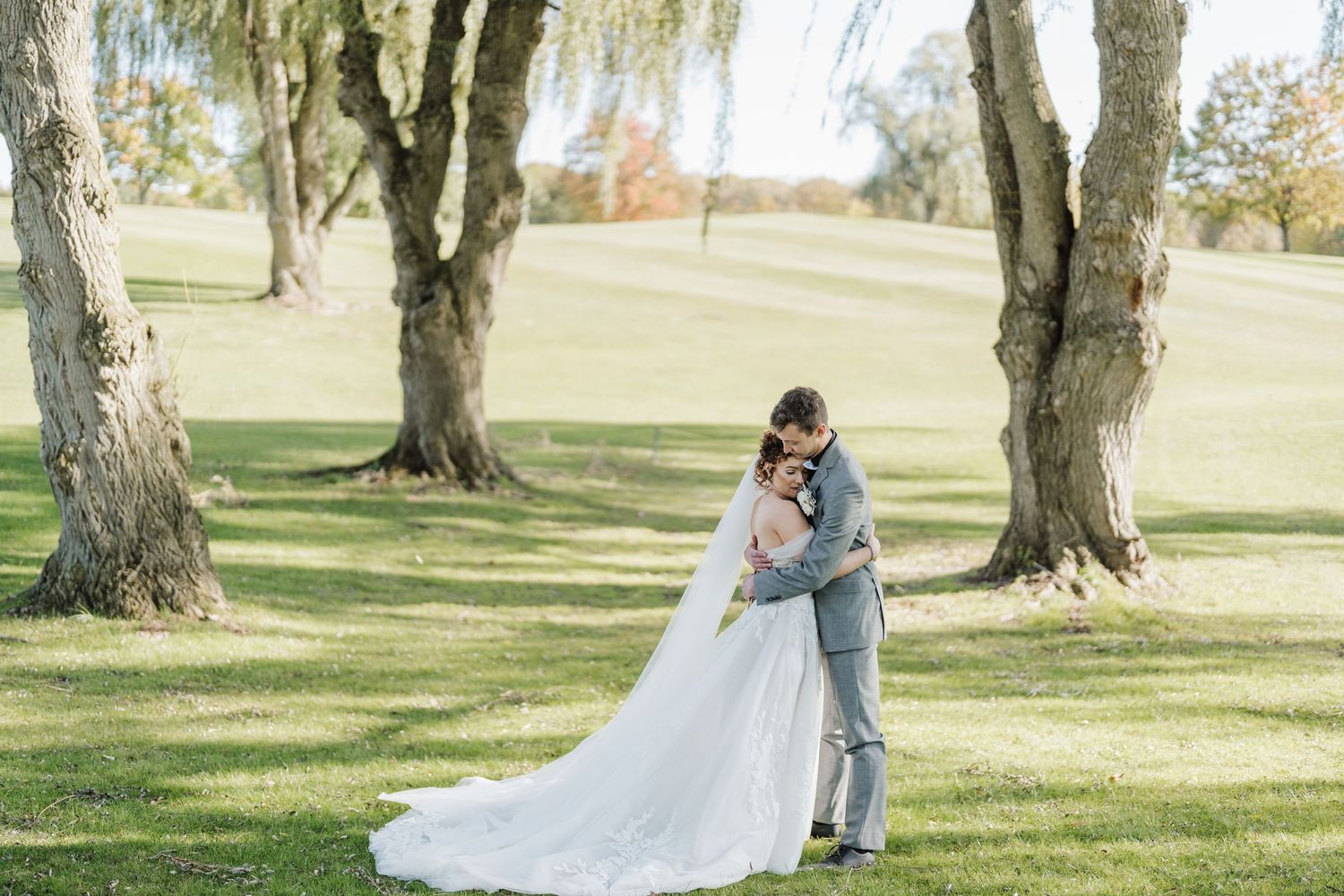 Couple in wedding attire embrace under weeping willow trees in a park setting.