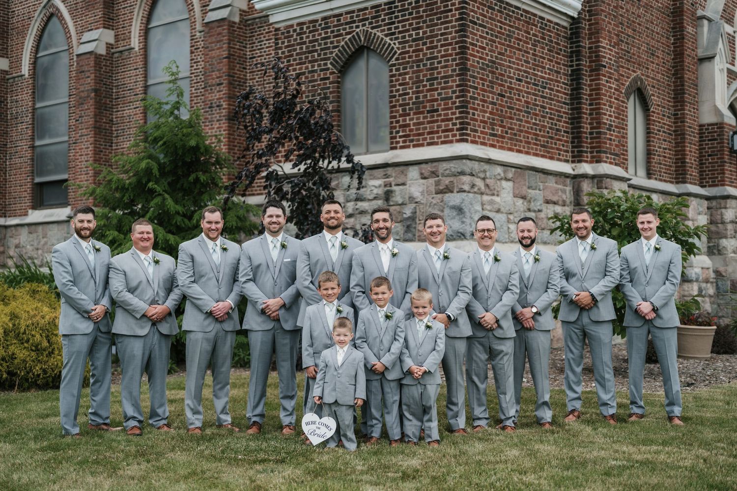 Groomsmen in matching grey suits pose with young ring bearers outside a brick church.