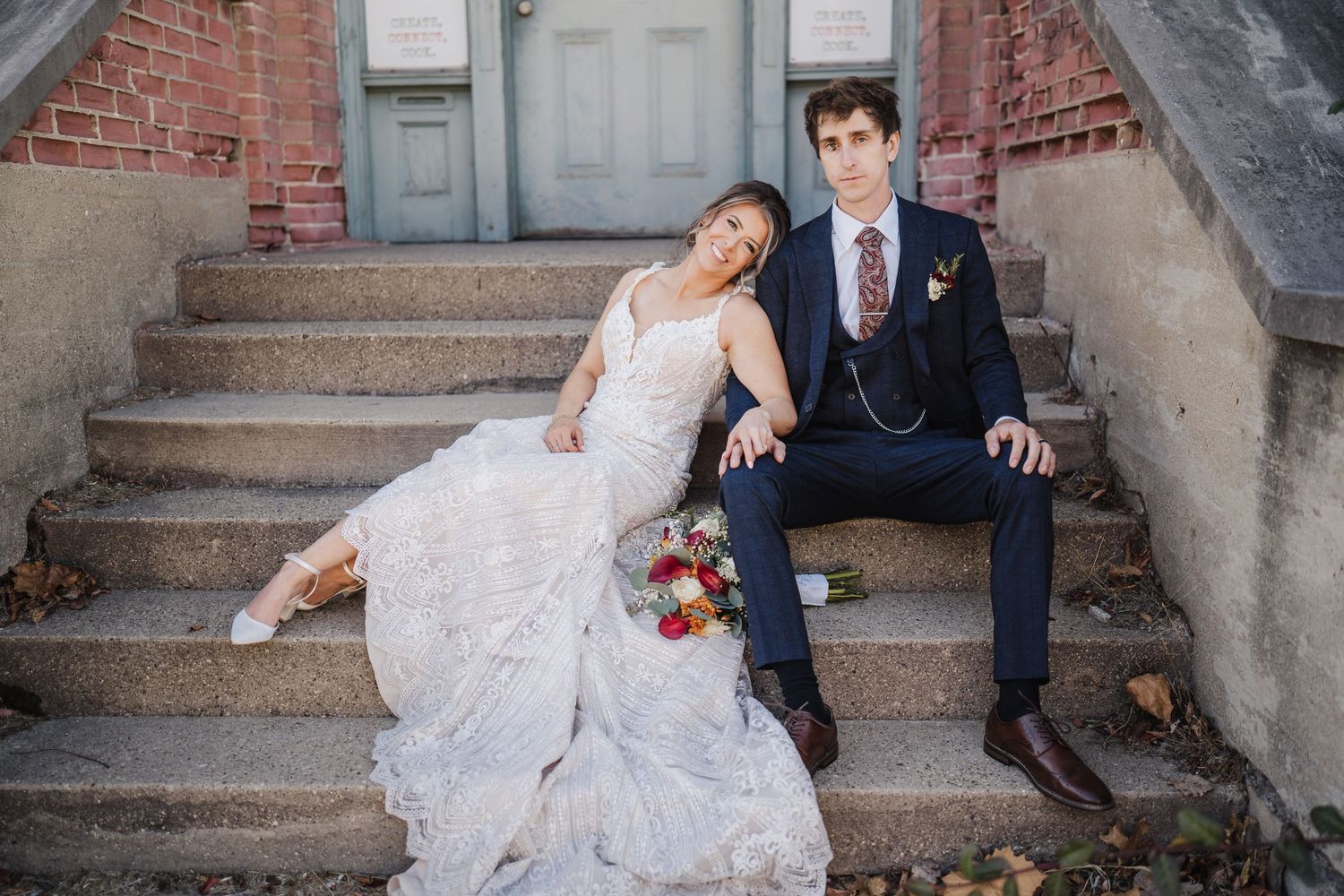 Couple poses casually on concrete steps in wedding attire with red brick wall backdrop.