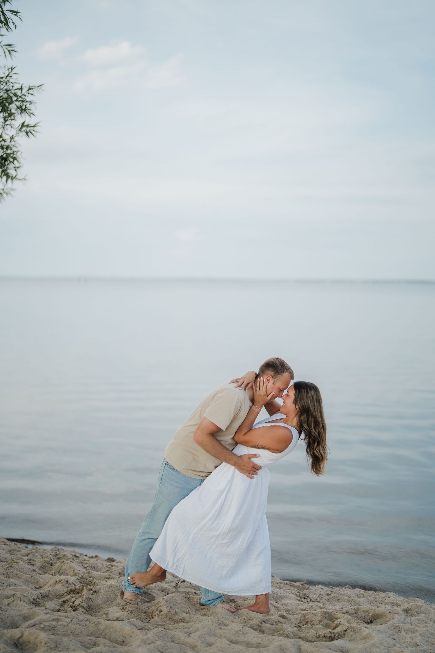 A romantic couple dances and embraces on a sandy beach at dusk with calm waters in the background.