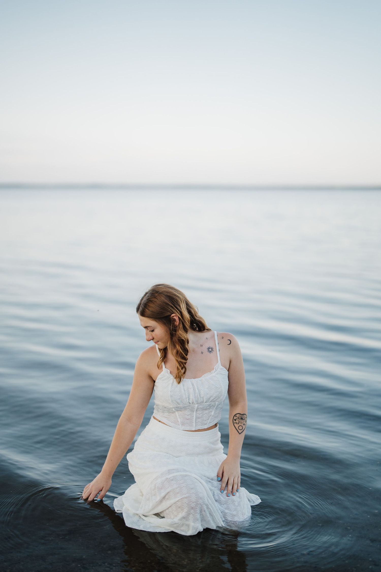 A figure in a white dress sits at the water's edge creating ripples in the calm blue water at sunset.