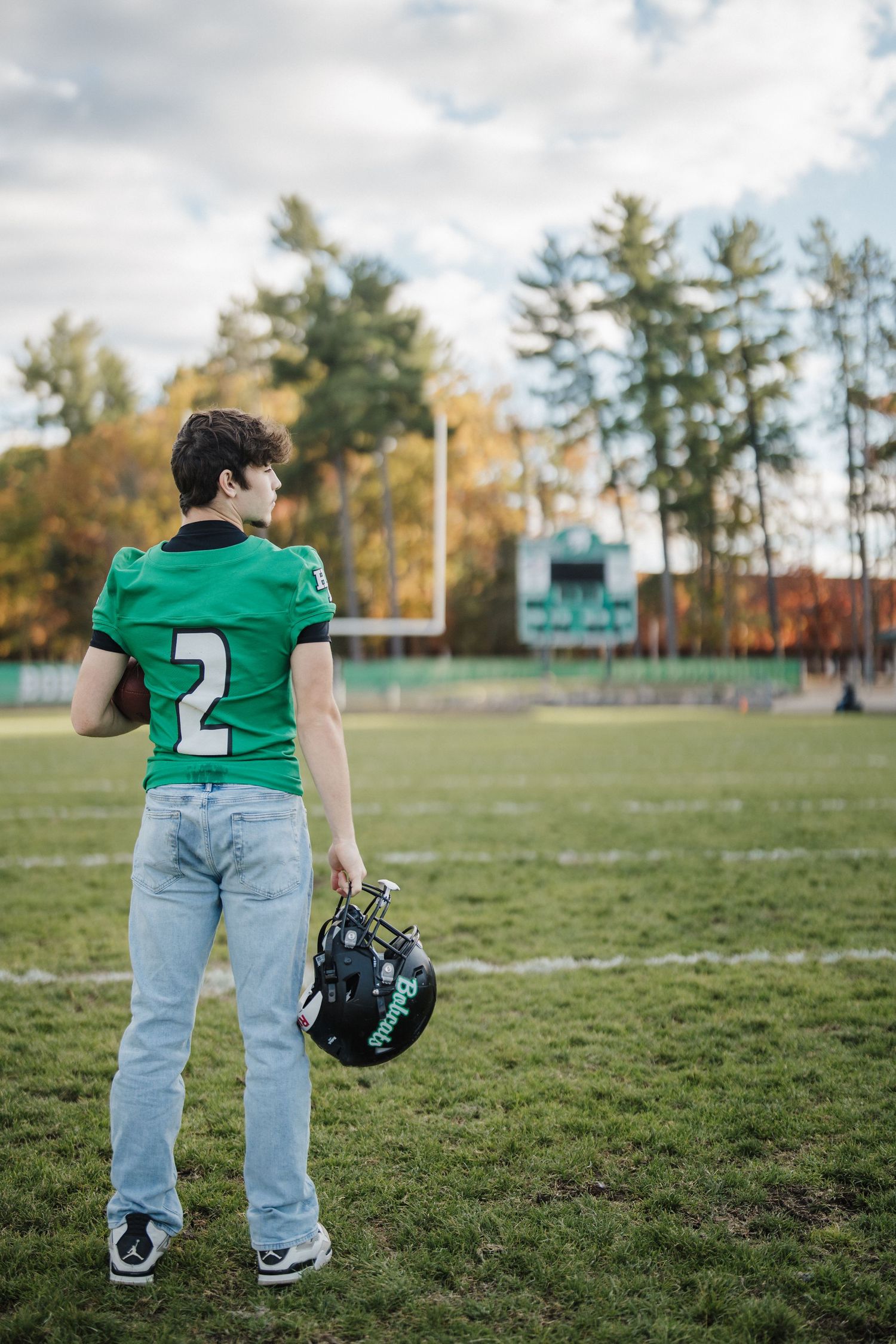 A person wearing a green jersey and jeans stands on a football field holding a helmet on an autumn day.