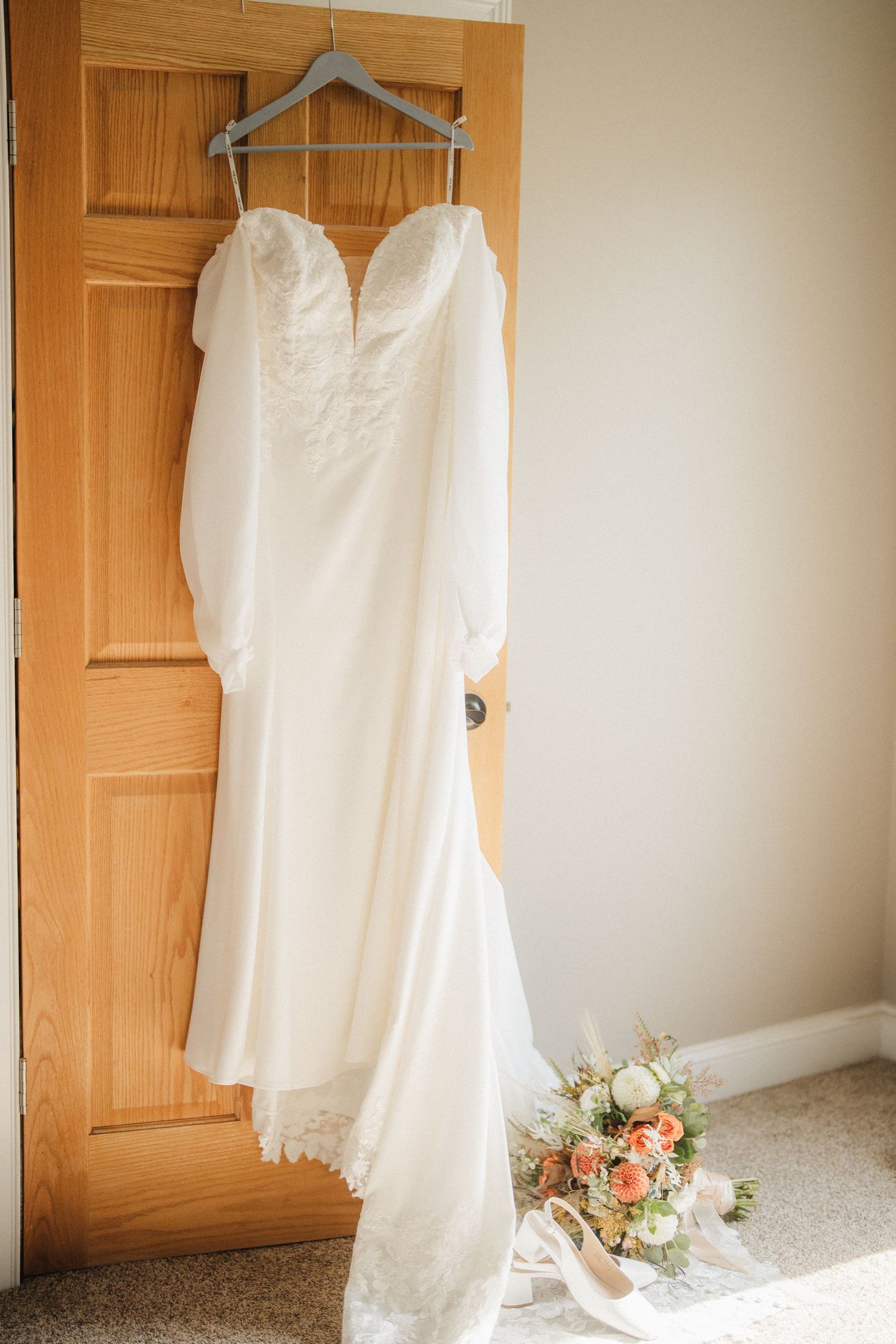 A white wedding dress hangs on a wooden door next to a bouquet of peach and white flowers.