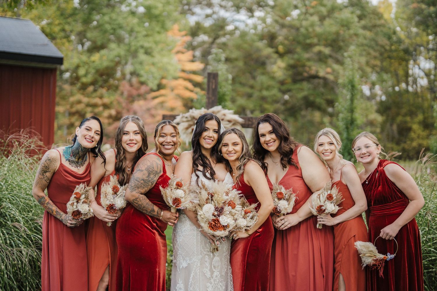 A group in red and burgundy dresses pose together holding bouquets during an autumn outdoor wedding celebration.