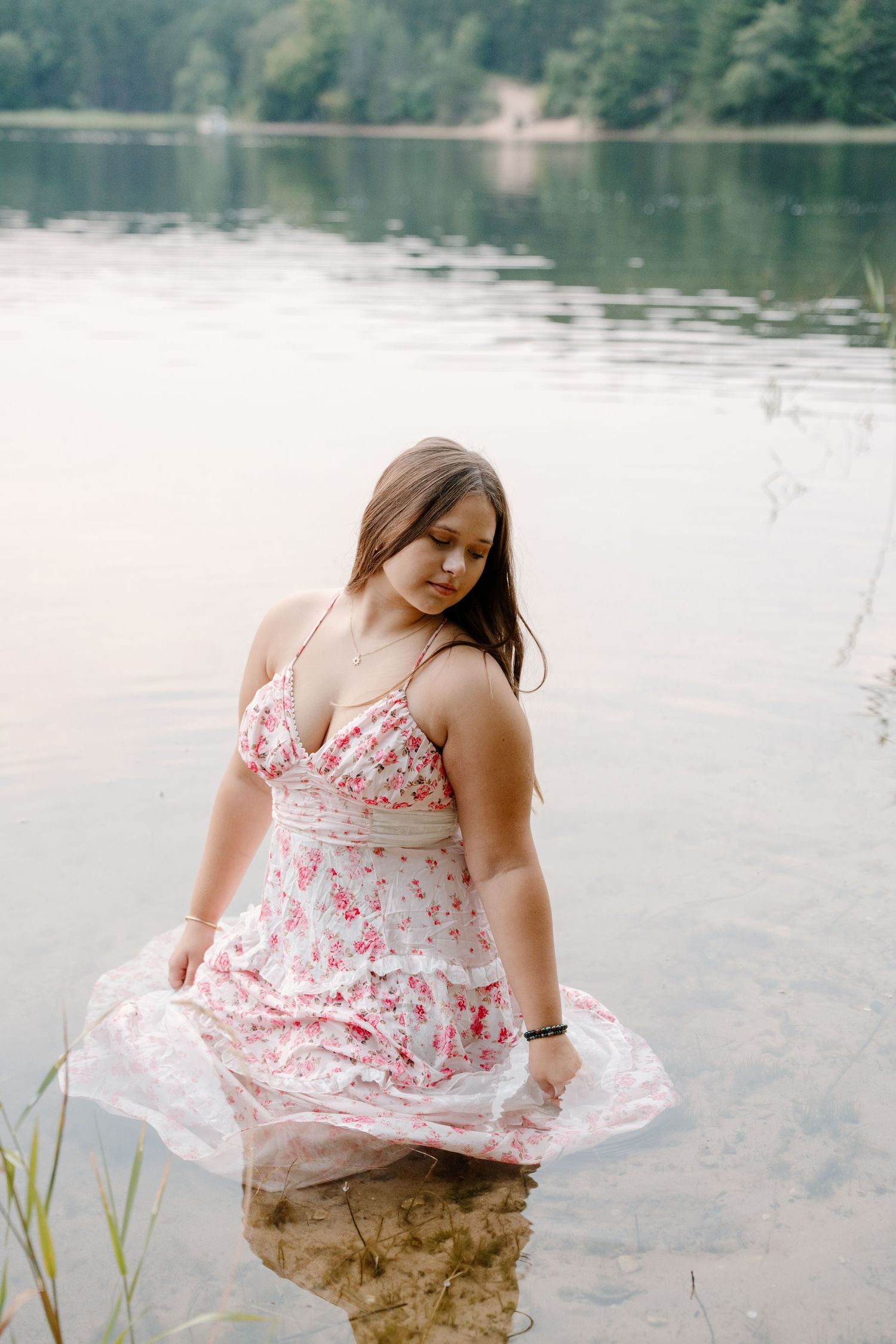 Young woman in floral sundress standing in calm lake water with lush green forest in background.