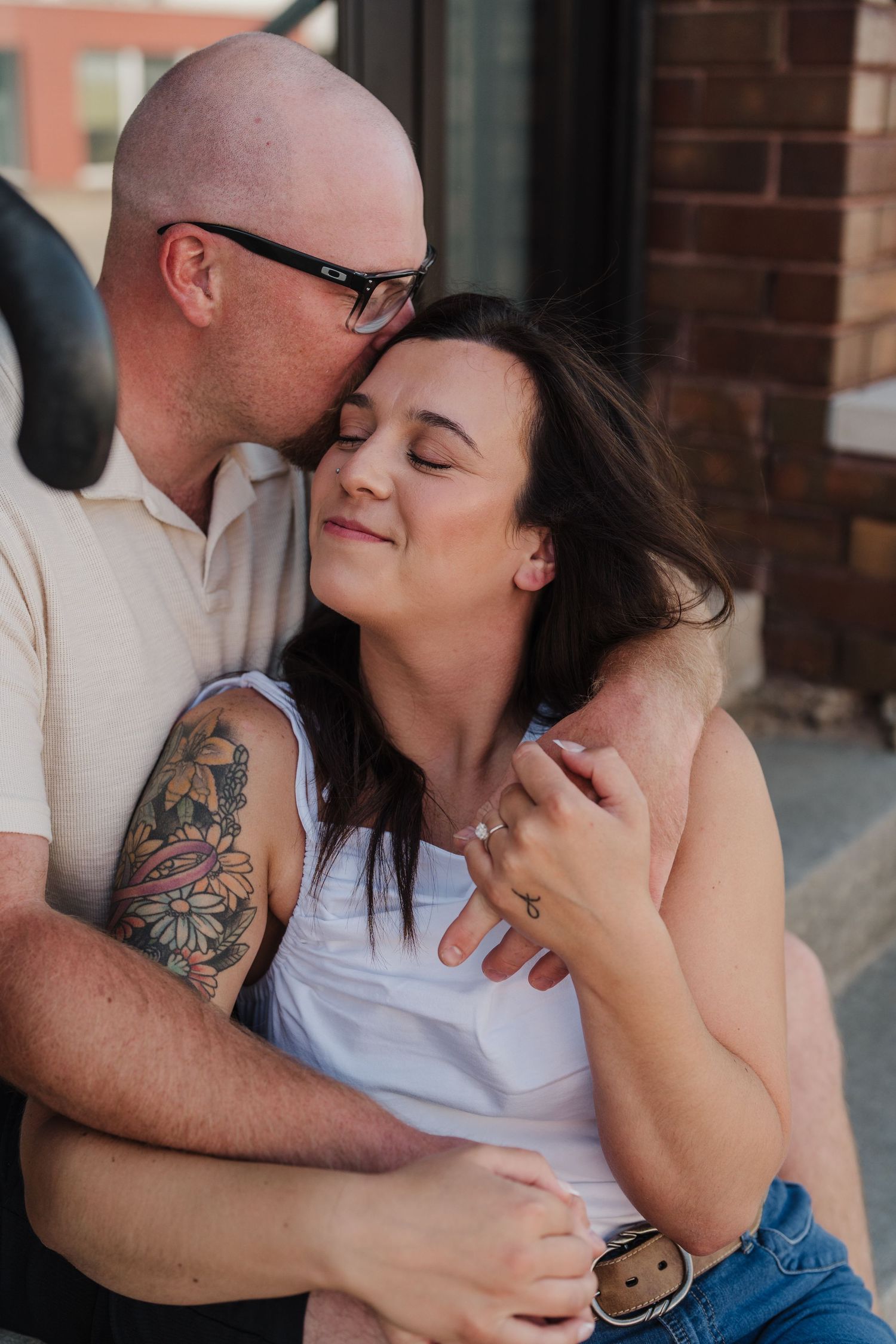 A bald man with glasses and tattoos kisses a dark-haired woman on the cheek as she smiles with eyes closed.