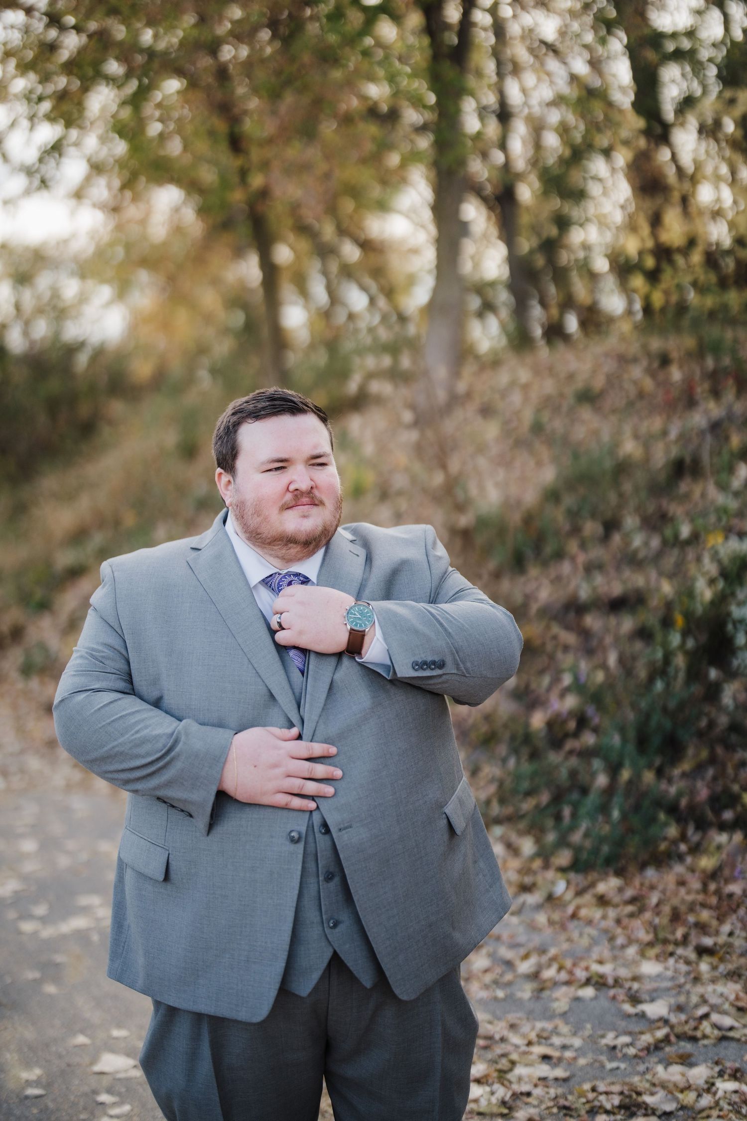 Groom in gray suit adjusts his tie while standing on a wooded path during autumn.