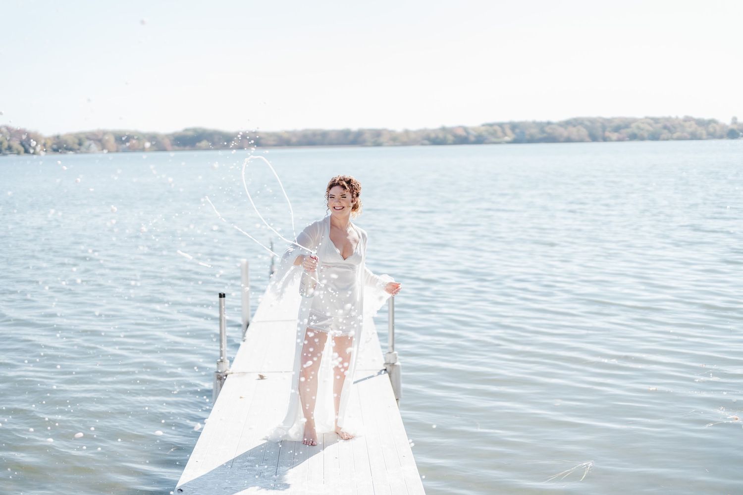 Person in a white dress stands on a dock by a serene lake on a bright summer day.