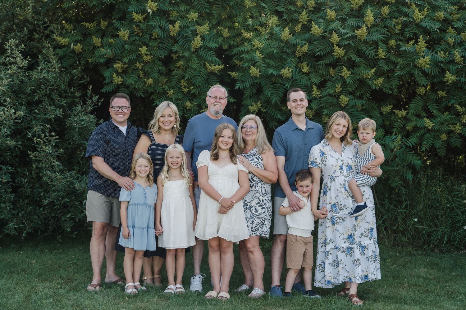 Large family group portrait smiling outdoors in front of lush green bushes on a sunny day.