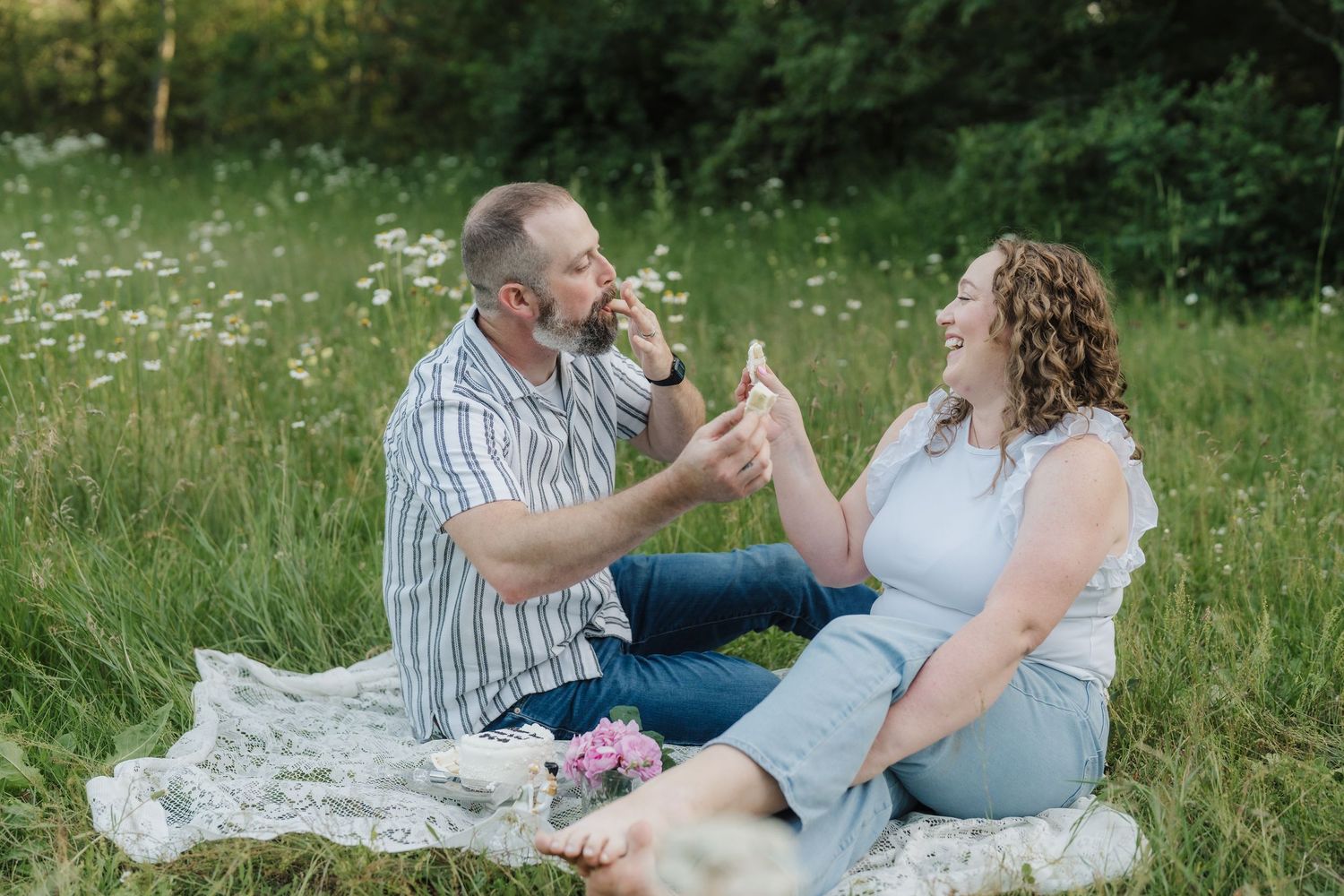 Couple laughing and sharing a drink during a romantic outdoor picnic in a green meadow with wildflowers.
