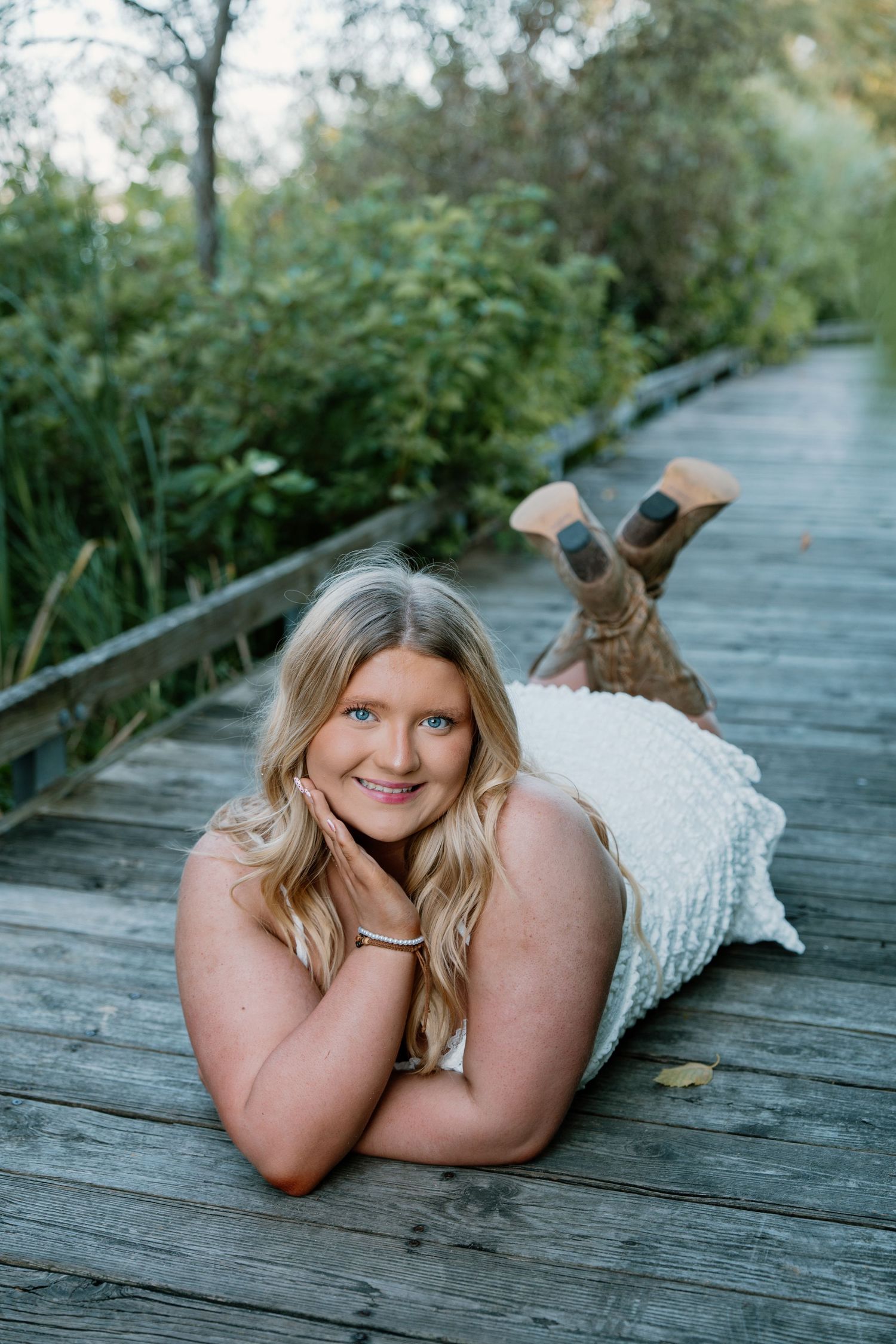 Smiling blonde woman lying on a wooden boardwalk surrounded by lush greenery, wearing a white dress and brown boots.