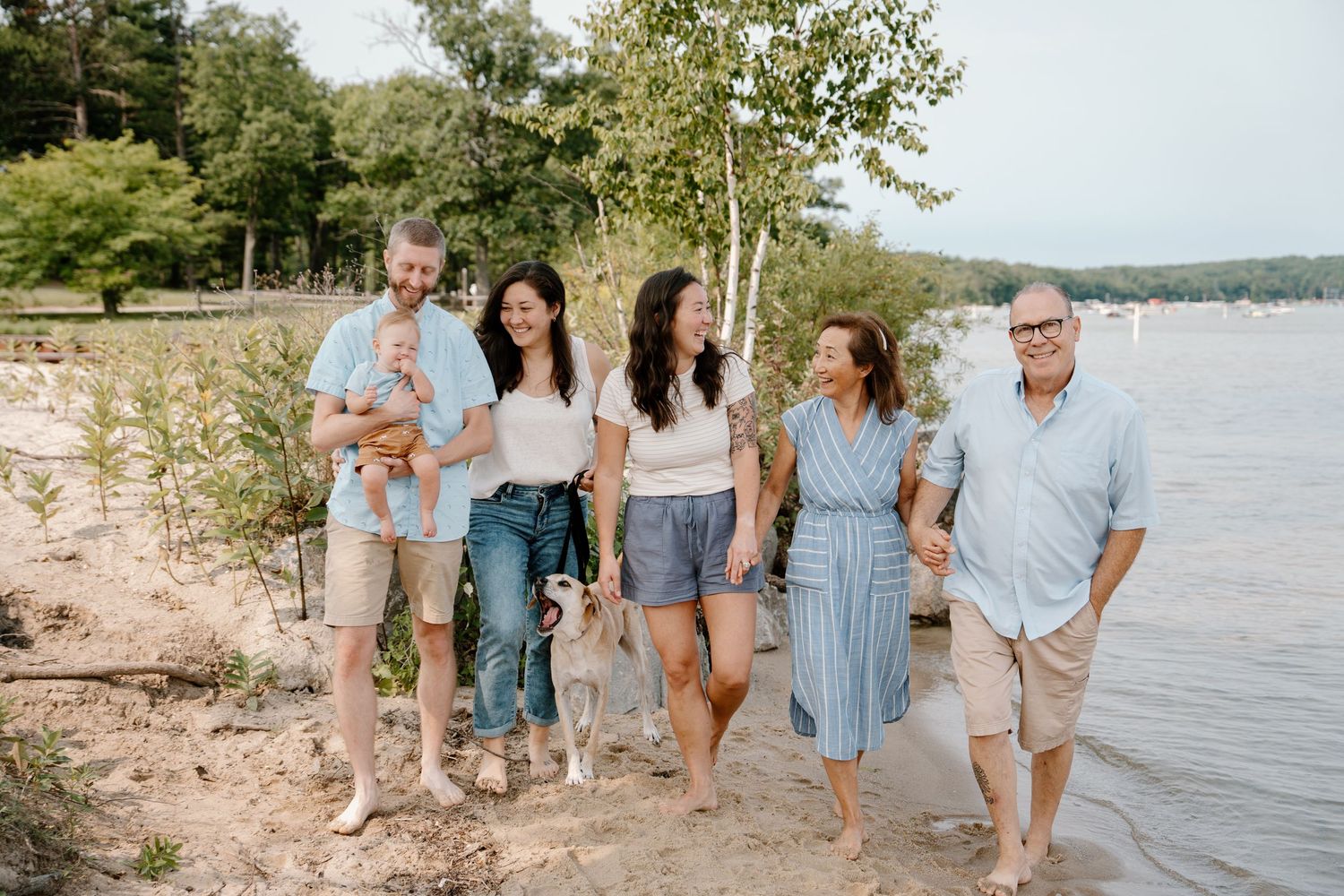 Family of six poses smiling on a sandy lakeside beach surrounded by trees on a warm summer day.