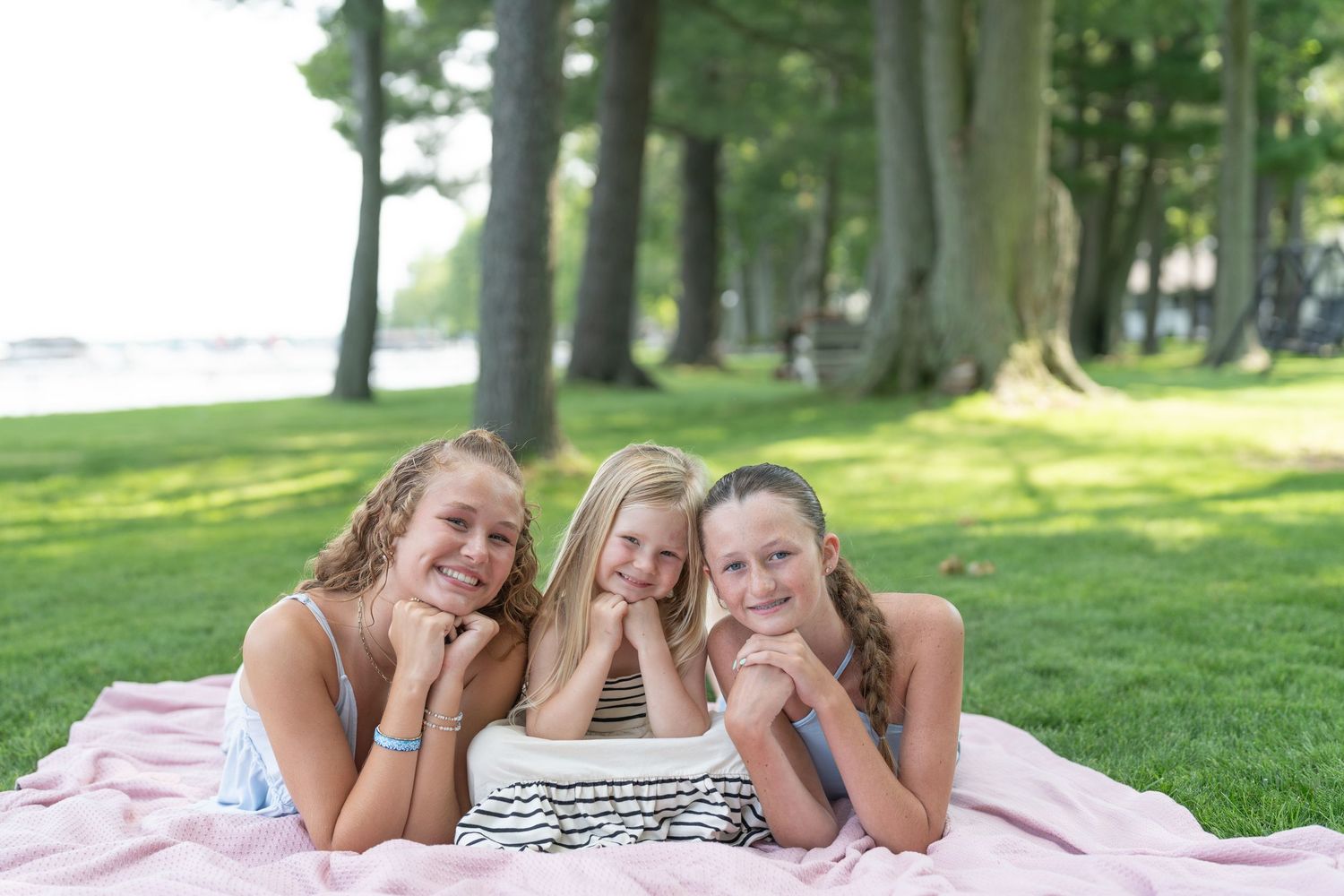 Three smiling girls lying on a pink blanket in a lush green park surrounded by tall trees.