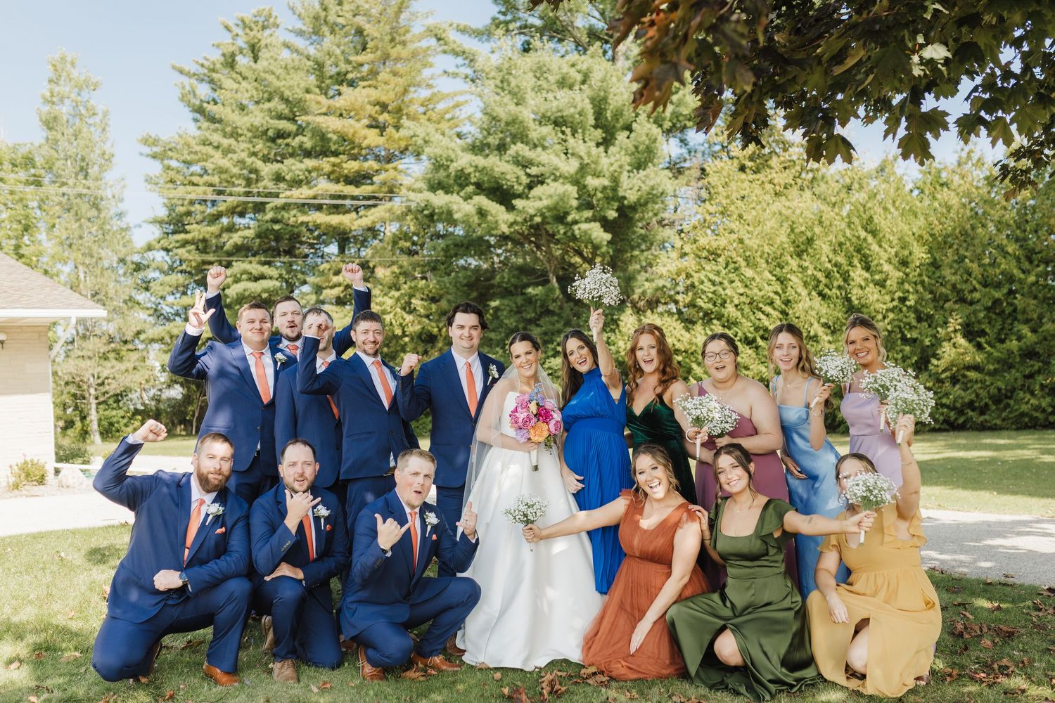 Wedding party in blue suits and colorful dresses pose together on a sunny lawn with trees in the background.
