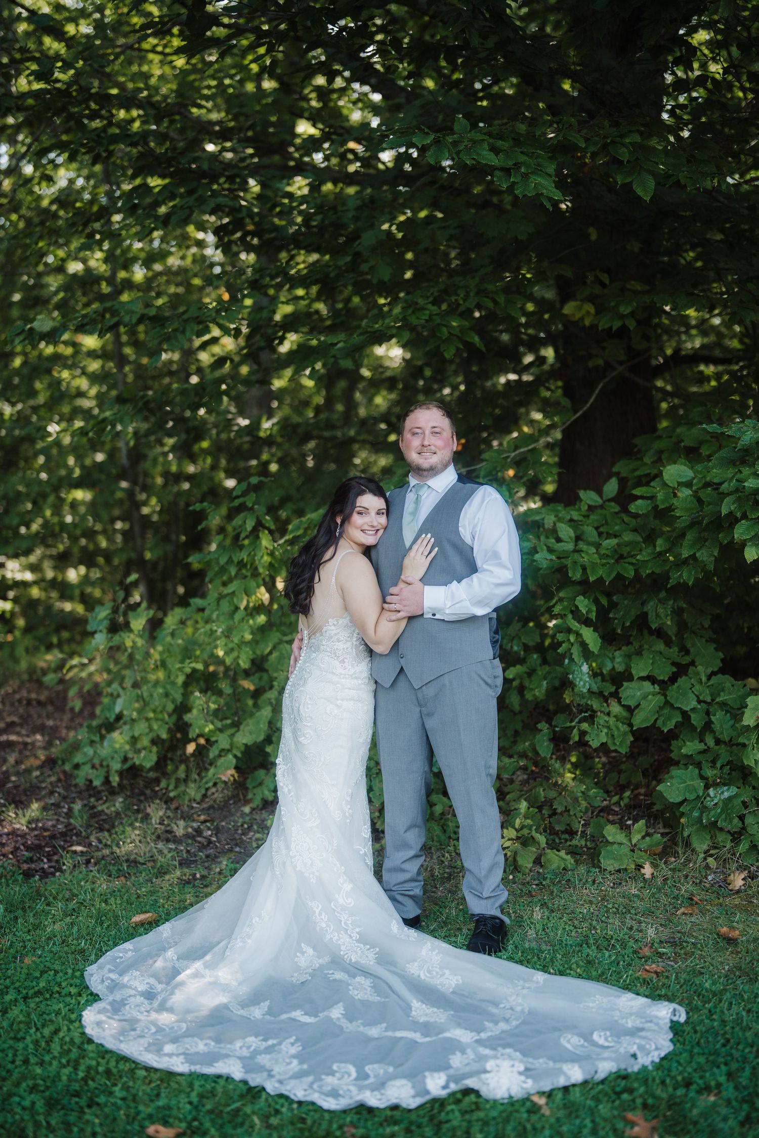 A couple in wedding attire poses outdoors in a natural garden setting with lush greenery and a flowing white lace dress.