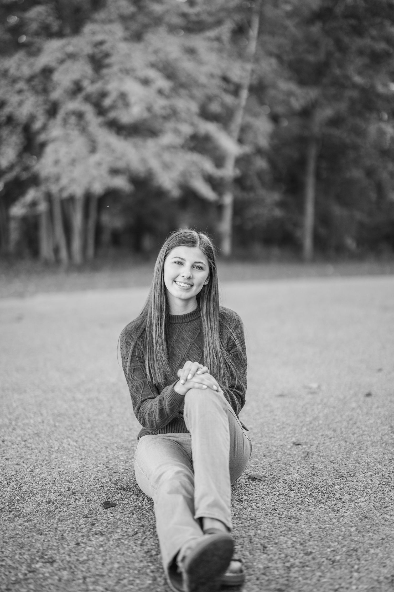Black and white portrait of someone sitting casually on a gravel path with trees in the background.