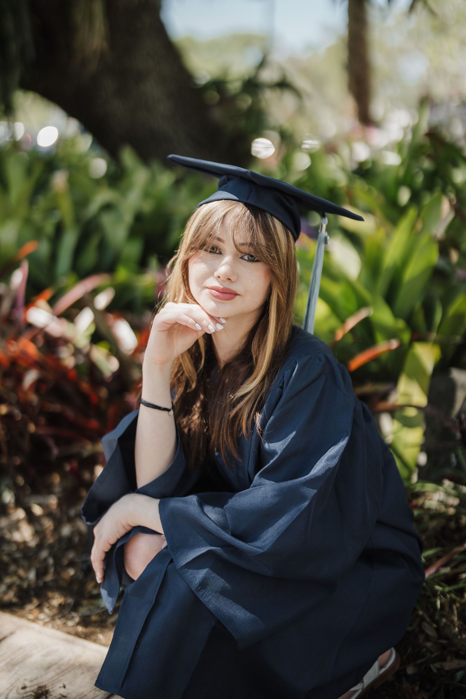 Graduate in navy blue academic regalia poses thoughtfully in a lush garden setting with natural sunlight.