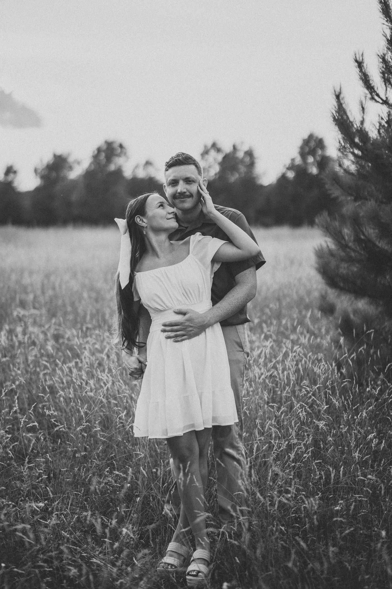 Two people in white dresses embrace while standing in a wildflower meadow at dusk.