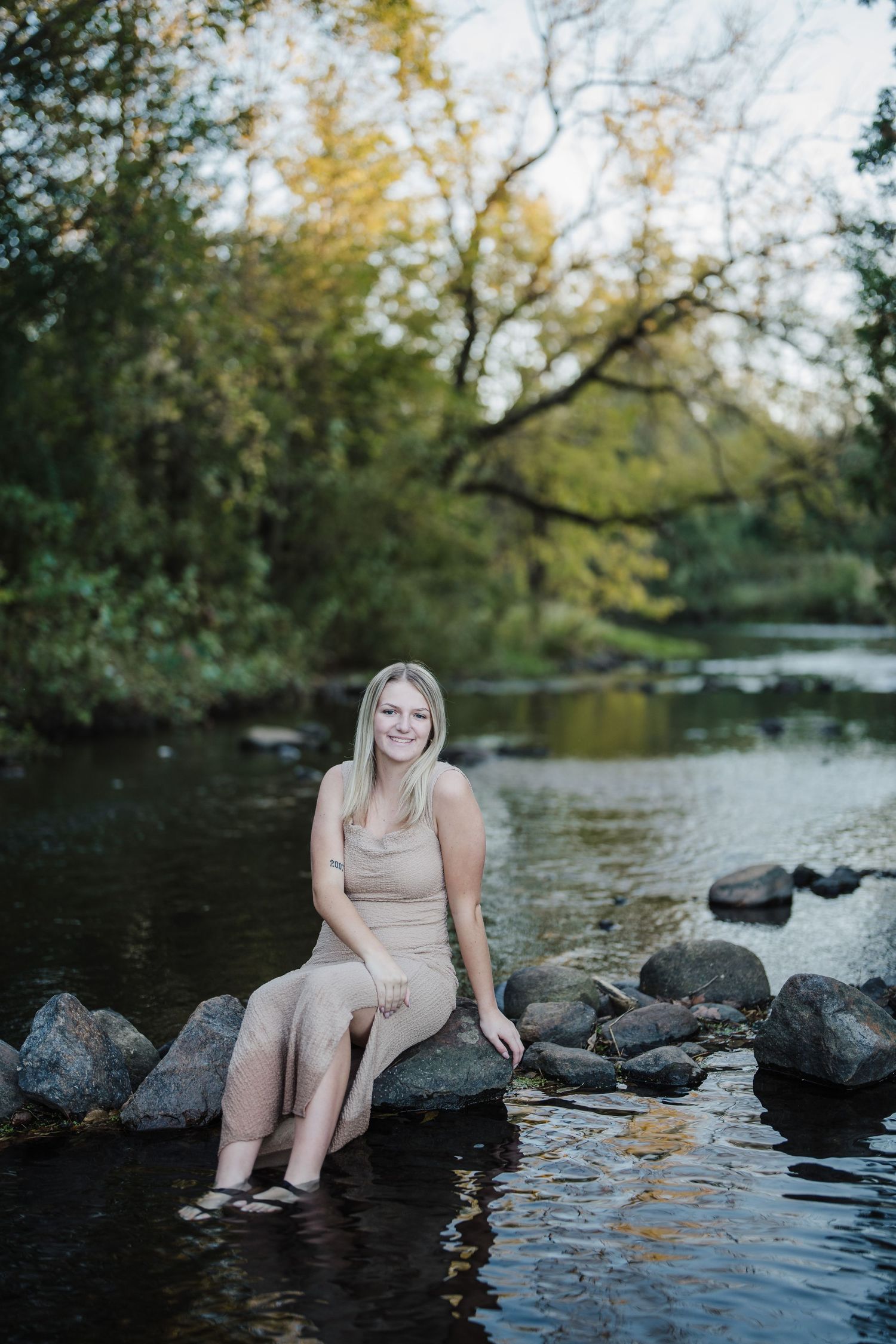 A person sits casually by a serene pond with rocks, surrounded by autumn trees and reflected water.