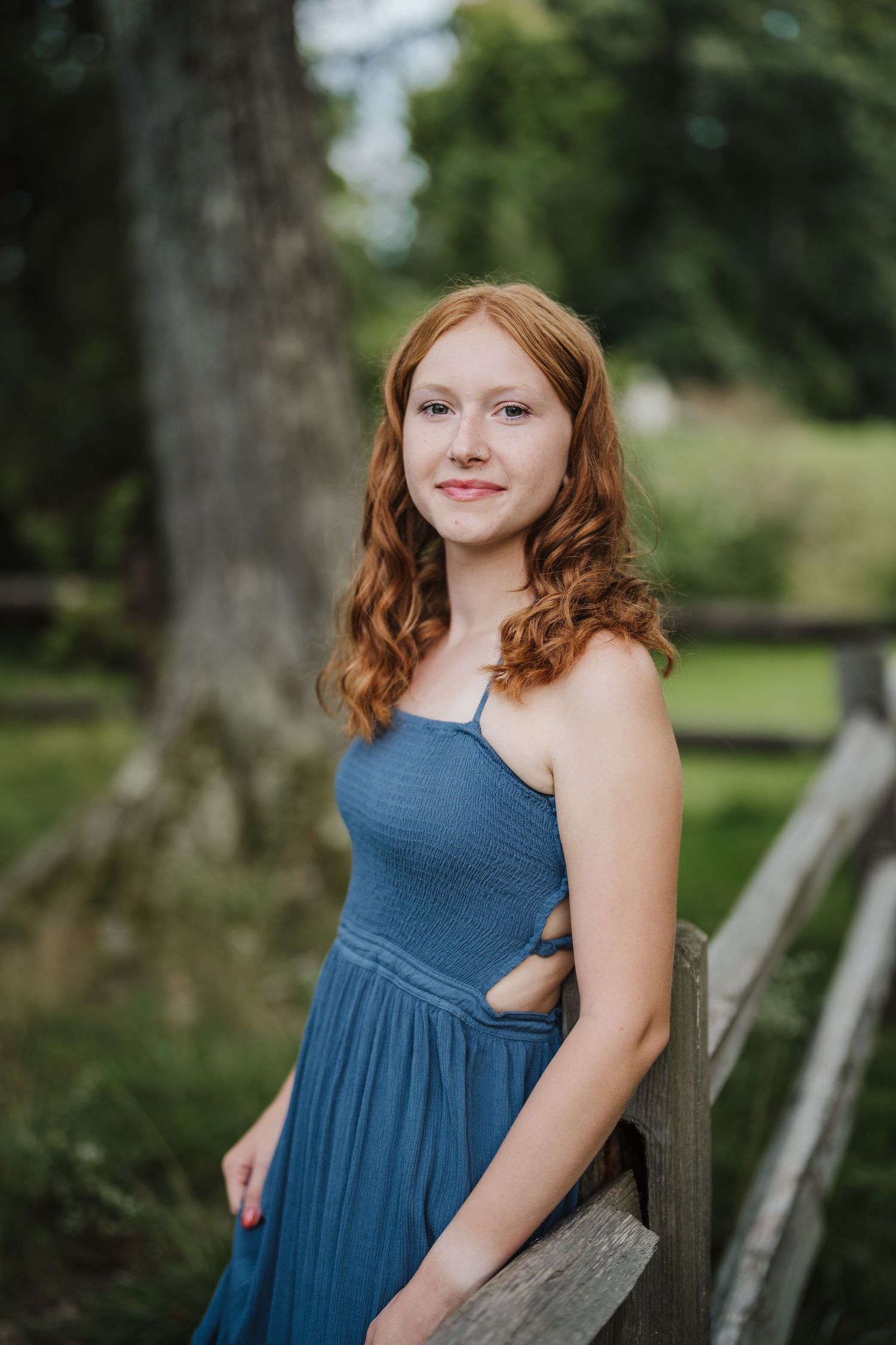 A person in a flowing blue dress poses near a rustic wooden fence in an outdoor garden setting.