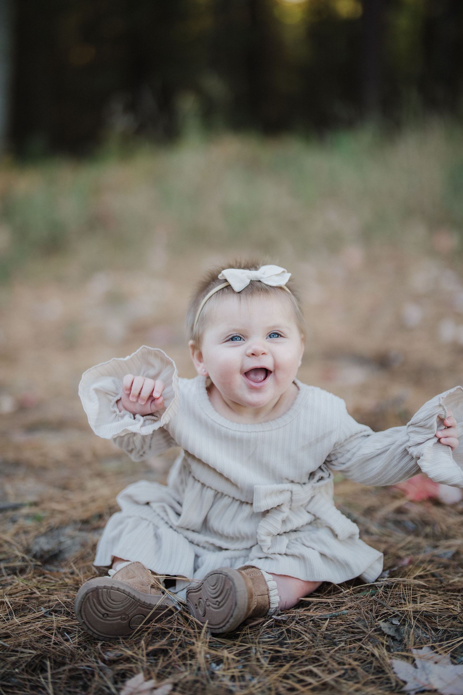 A baby in a cream-colored sweater dress sits on fallen autumn leaves while smiling and playing outdoors.