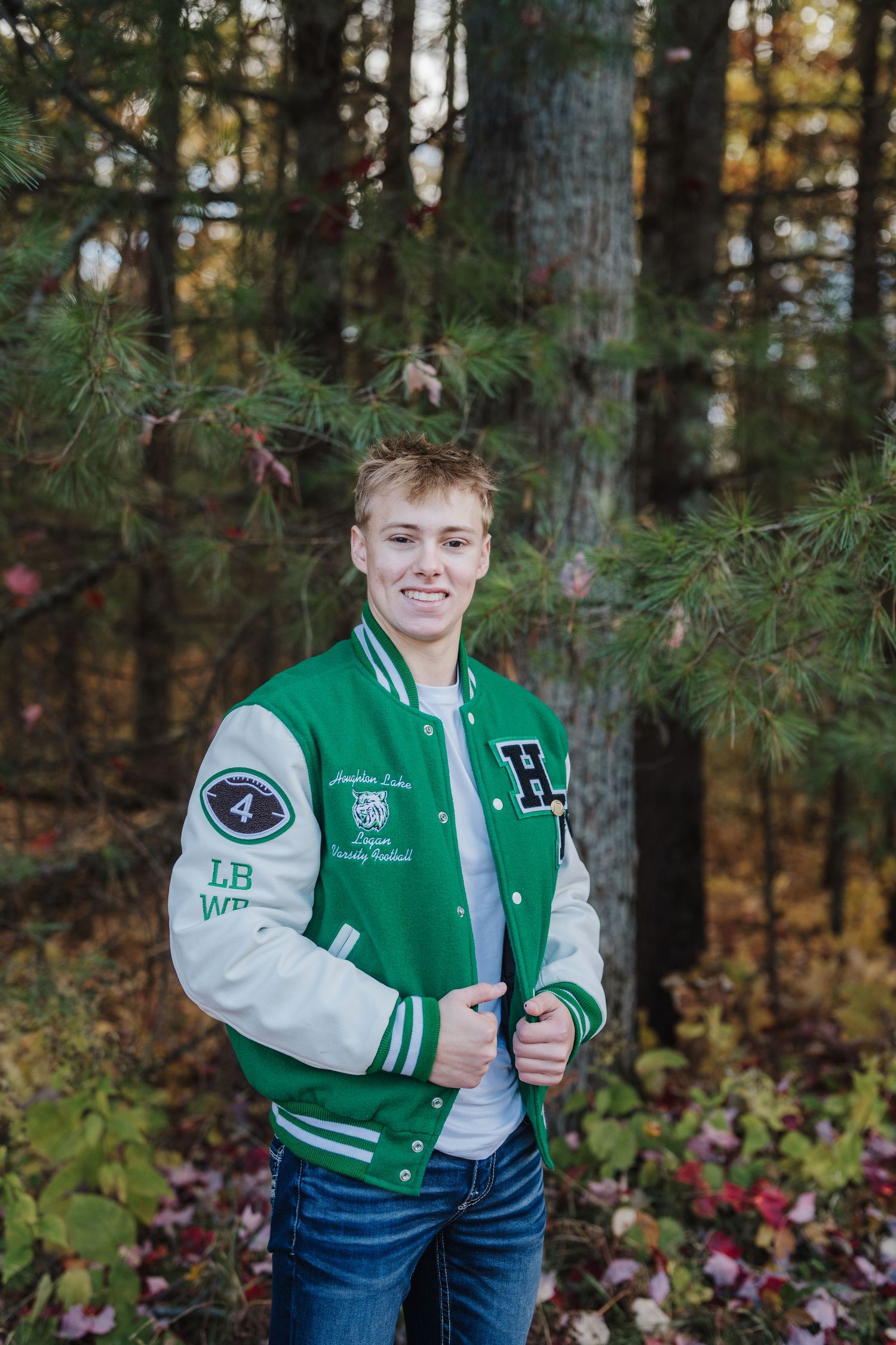 Student in varsity letterman jacket poses for portrait in wooded outdoor setting.