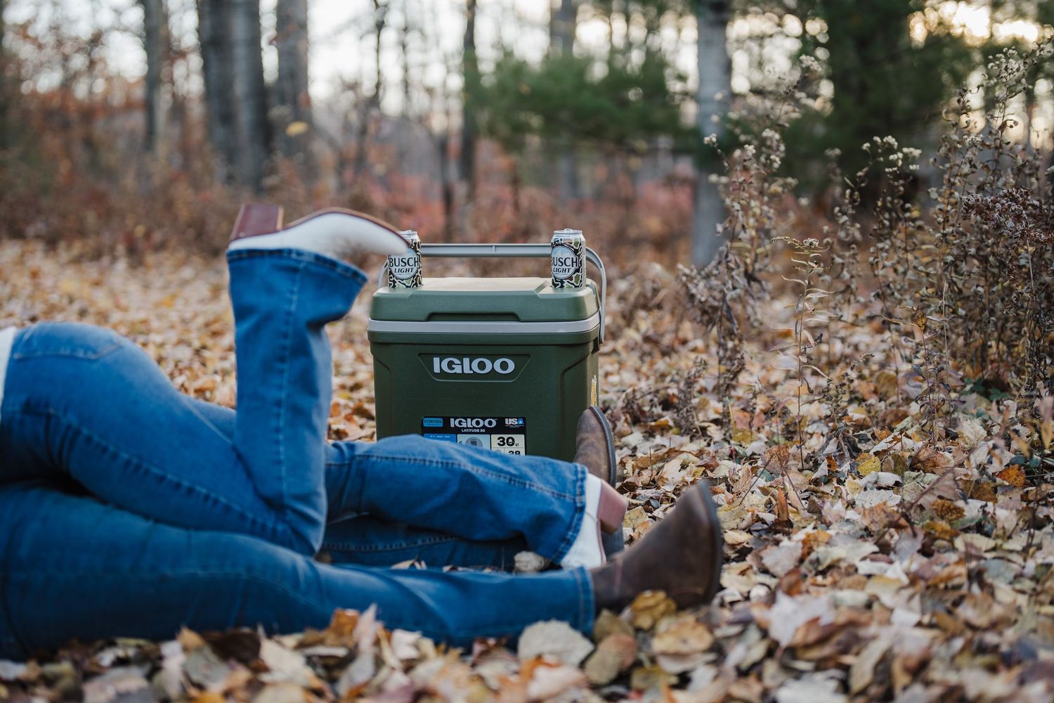 Cozy fall scene with someone wearing blue jeans relaxing on fallen leaves while holding a thermos outdoors.