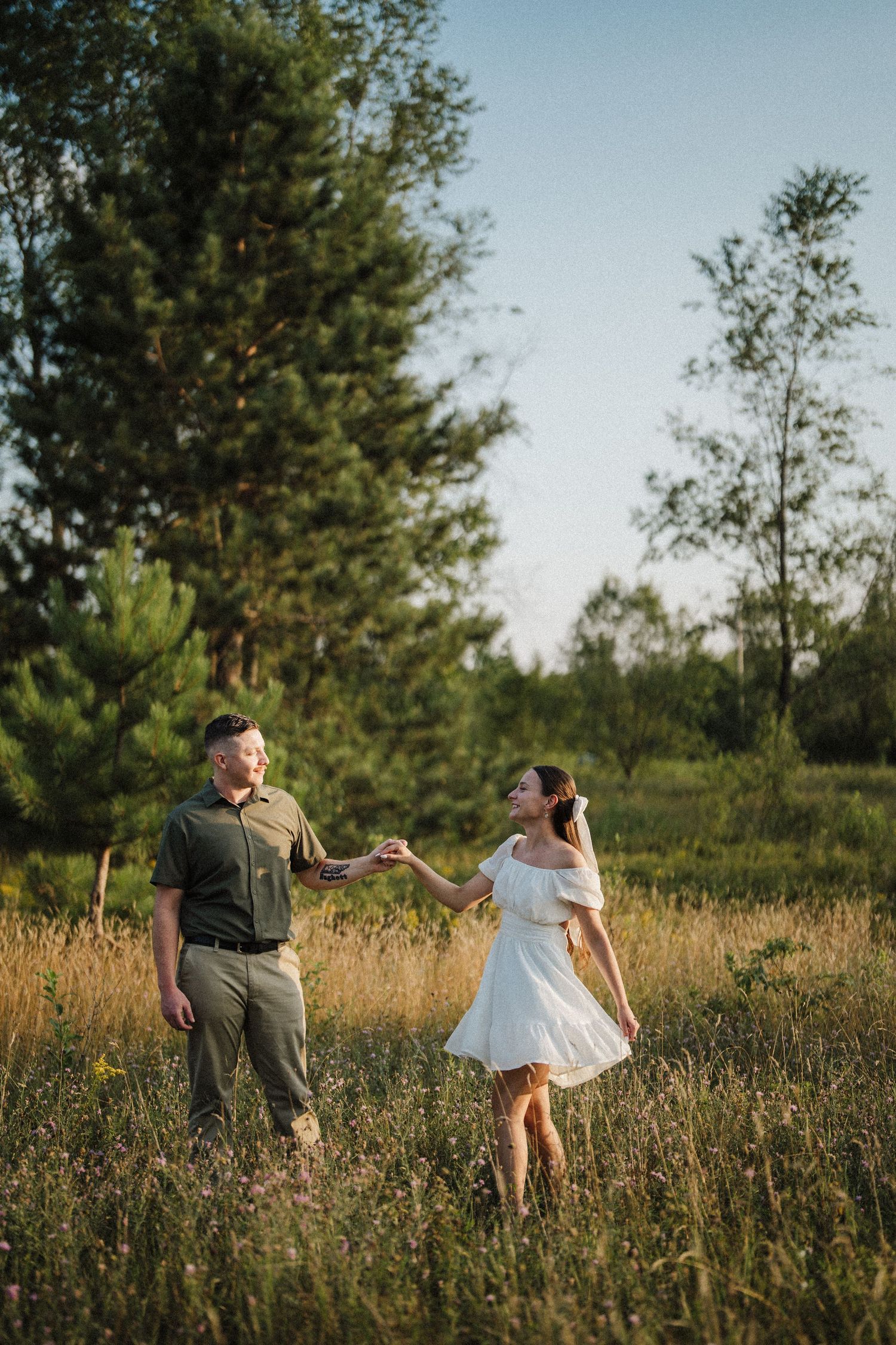 A couple holds hands in a golden meadow at sunset with pine trees in the background.