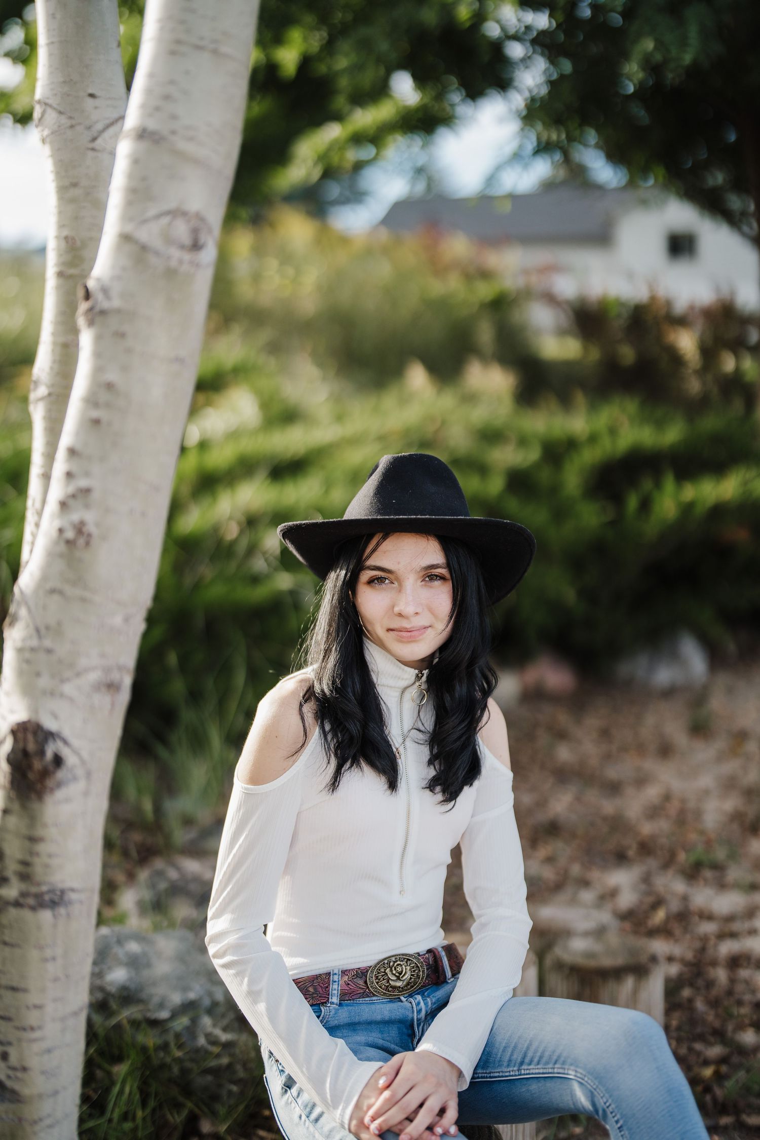 A person in a white blouse and black hat poses against white birch trees in natural outdoor lighting.