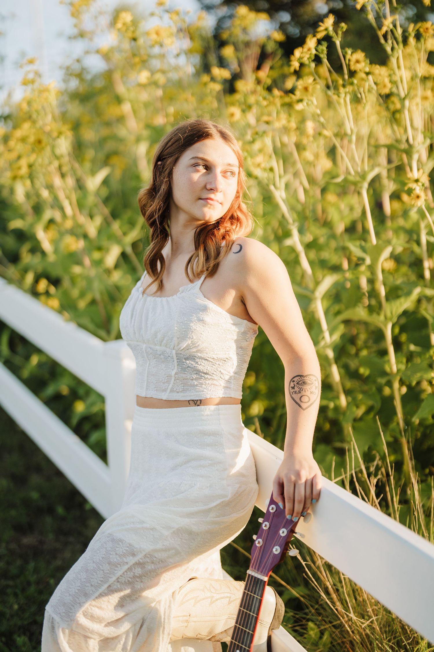Sunlit portrait with white lace outfit and guitar against rustic fence in golden hour.
