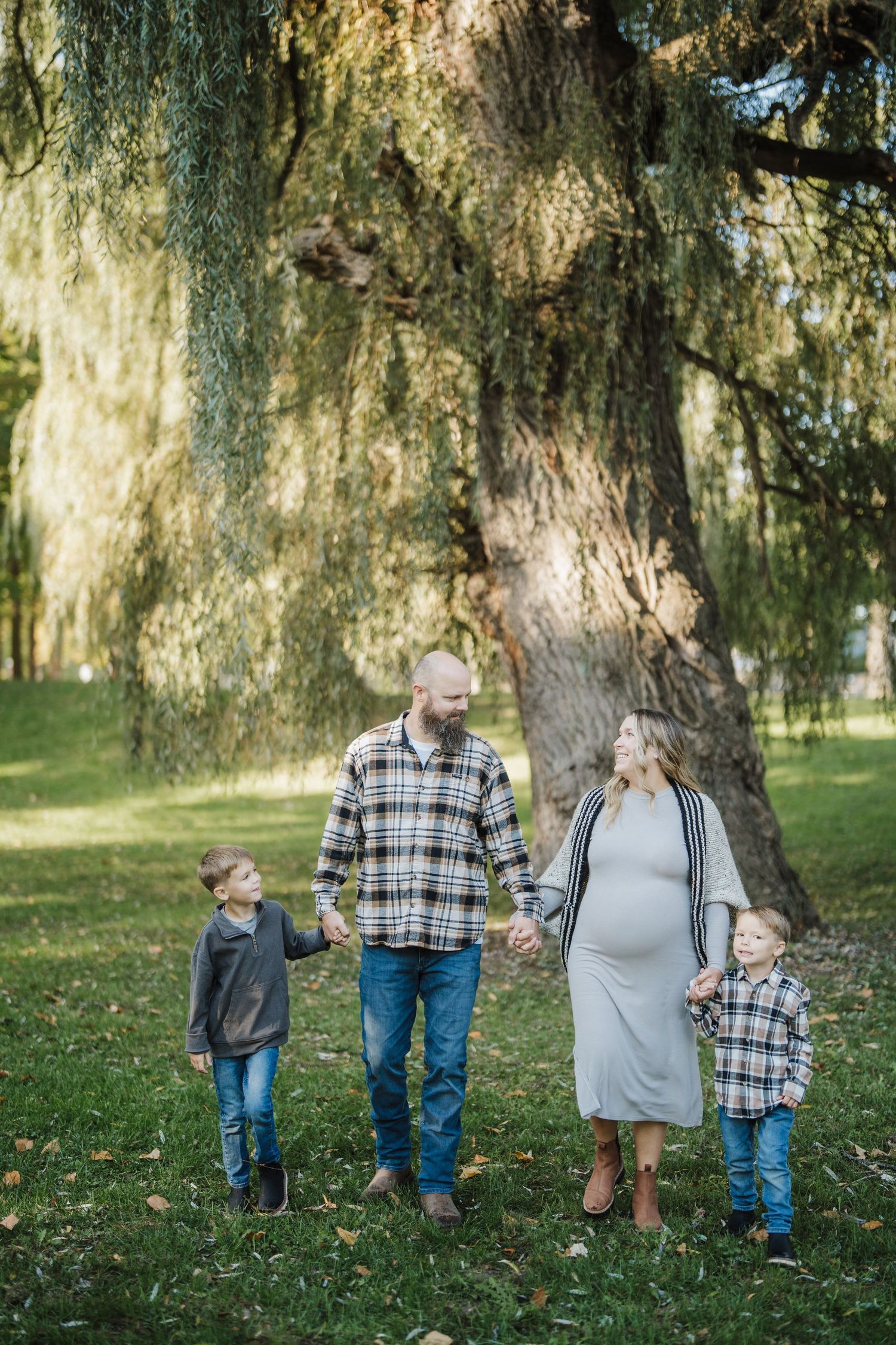 A family of three stands together under a large weeping willow tree in a peaceful park setting.