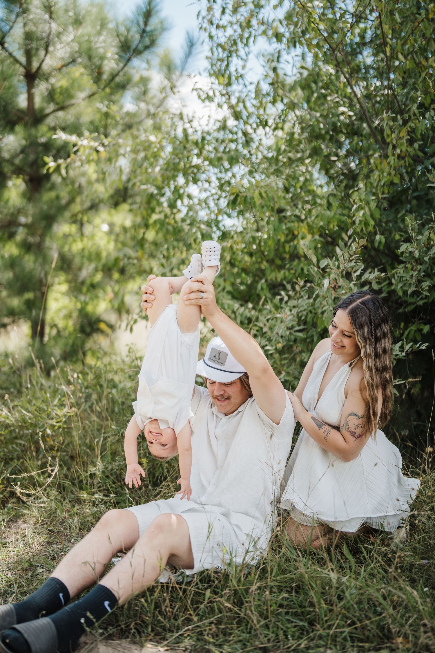 A family shares joyful moments while relaxing on grass beneath trees in coordinated white outfits.