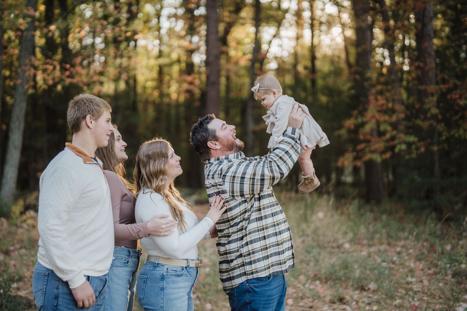 A family in coordinated casual outfits laughs together during an outdoor autumn photo session in a wooded area.